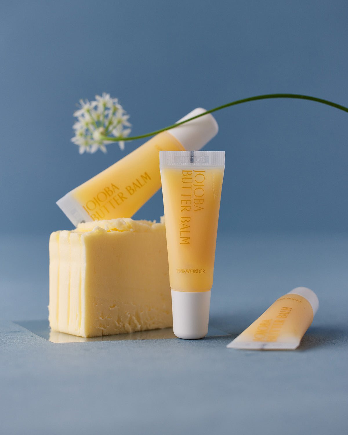 An artfully styled image on a blue background featuring a cube of butter. Three small yellow tubes of Jojoba Butter Balm are placed around it, along with a white, delicate flower on a thin green stem, playing on the "butter balm" name.