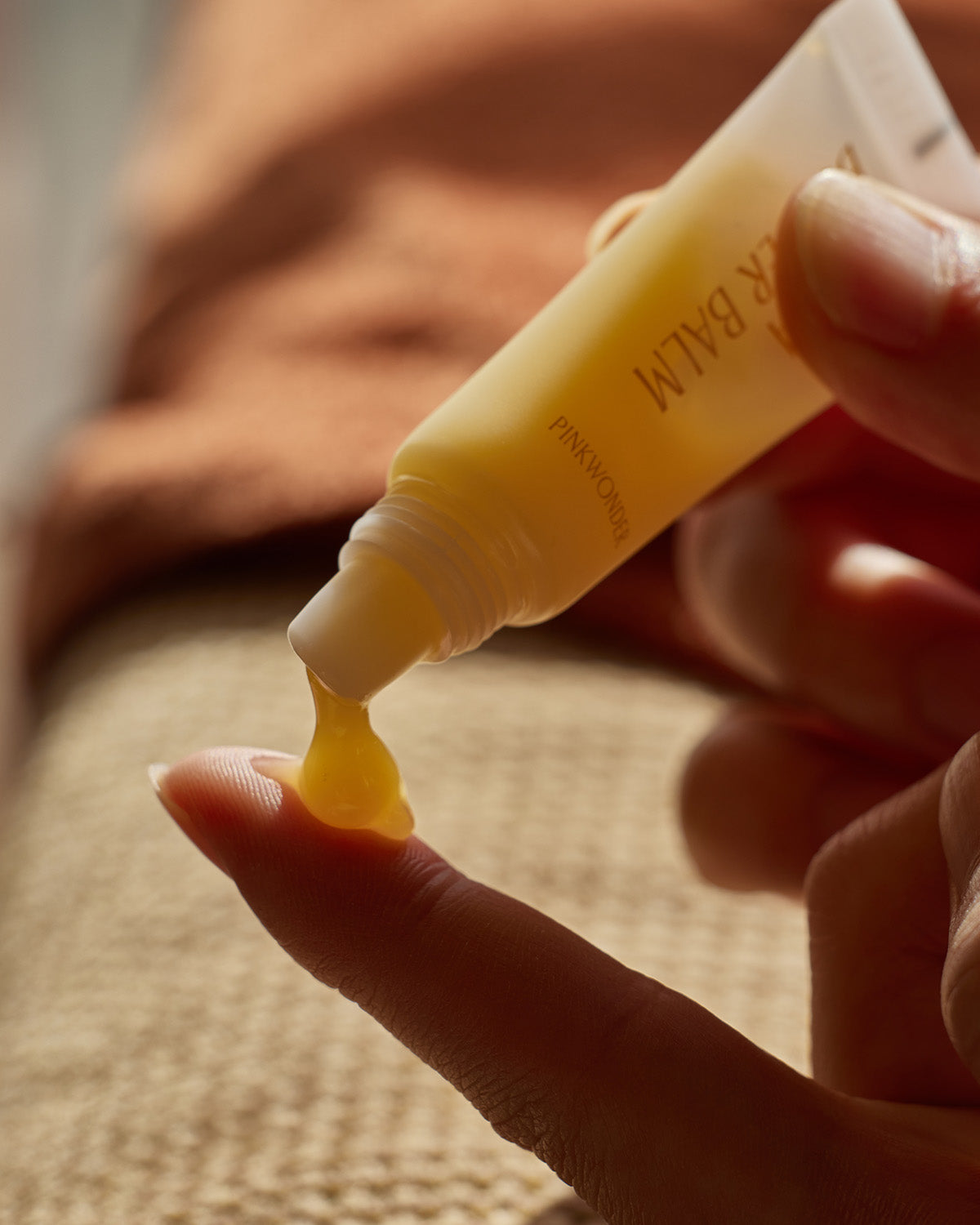 A close-up action shot of a person's fingers dispensing a bead of thick, translucent, yellow-gold Jojoba Butter Balm from the small tube onto the tip of their finger. The background is warm and blurred.