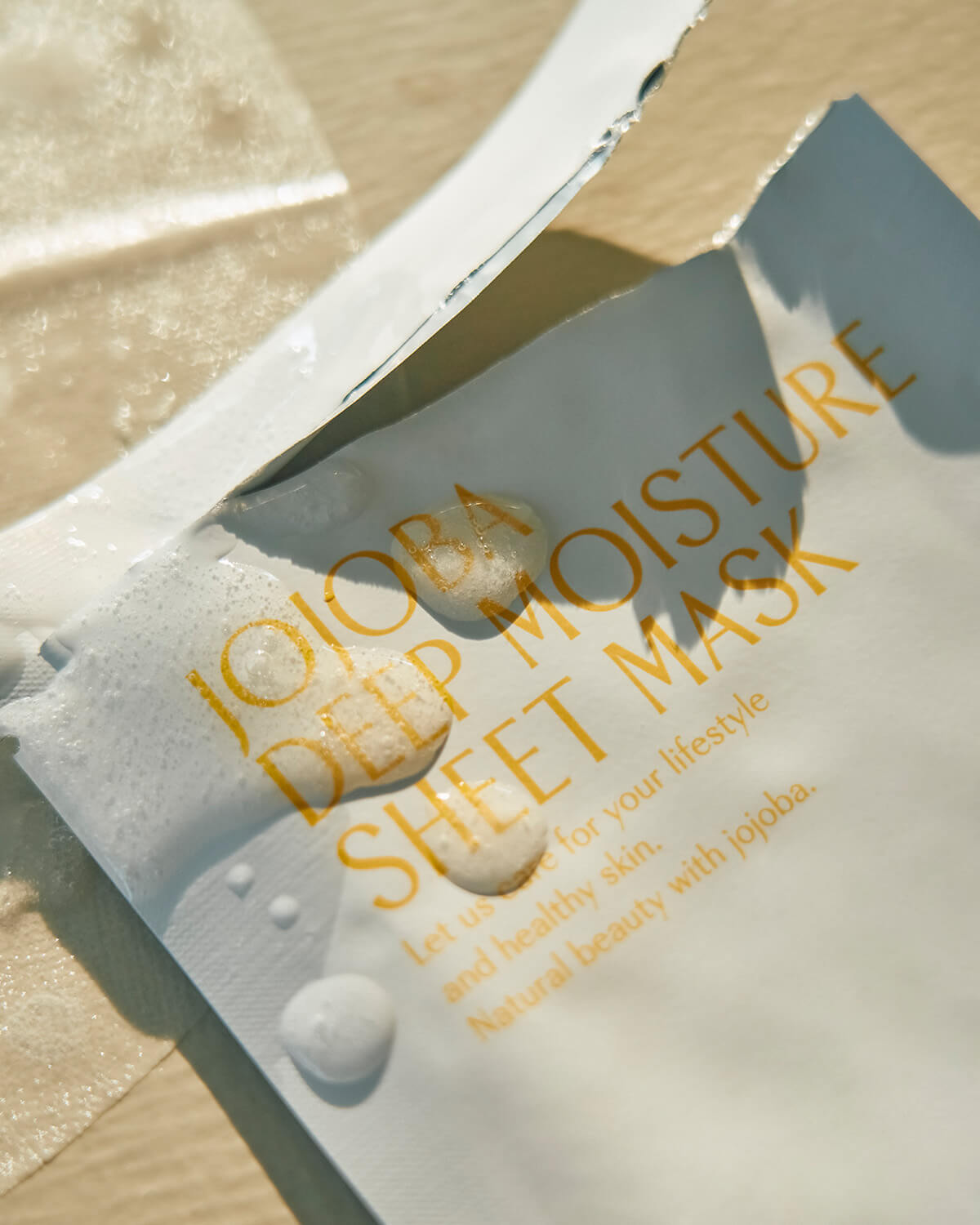 A close-up of the opened white foil packet for the Jojoba Deep Moisture Sheet Mask. Several large drops of clear liquid serum are resting on the yellow text of the mask packaging.