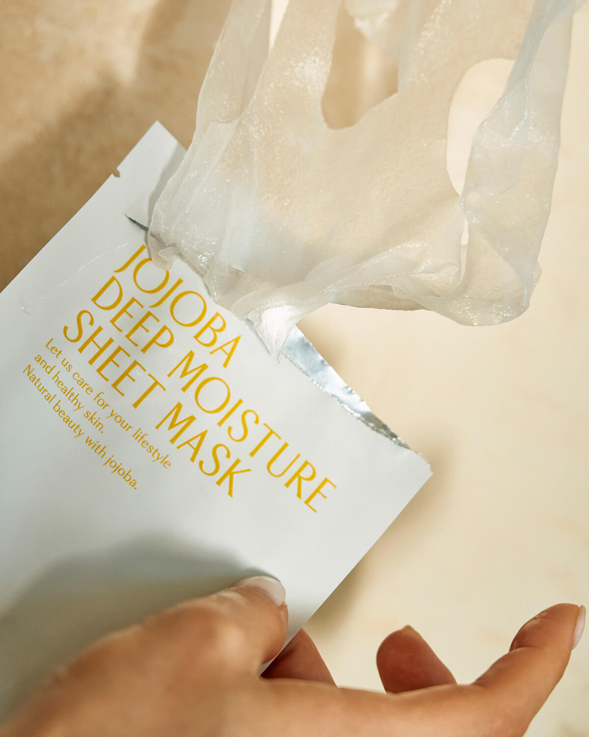 A close-up, overhead shot of a person's hand opening the white foil packet for the Jojoba Deep Moisture Sheet Mask, revealing the drenched, translucent mask being pulled out.