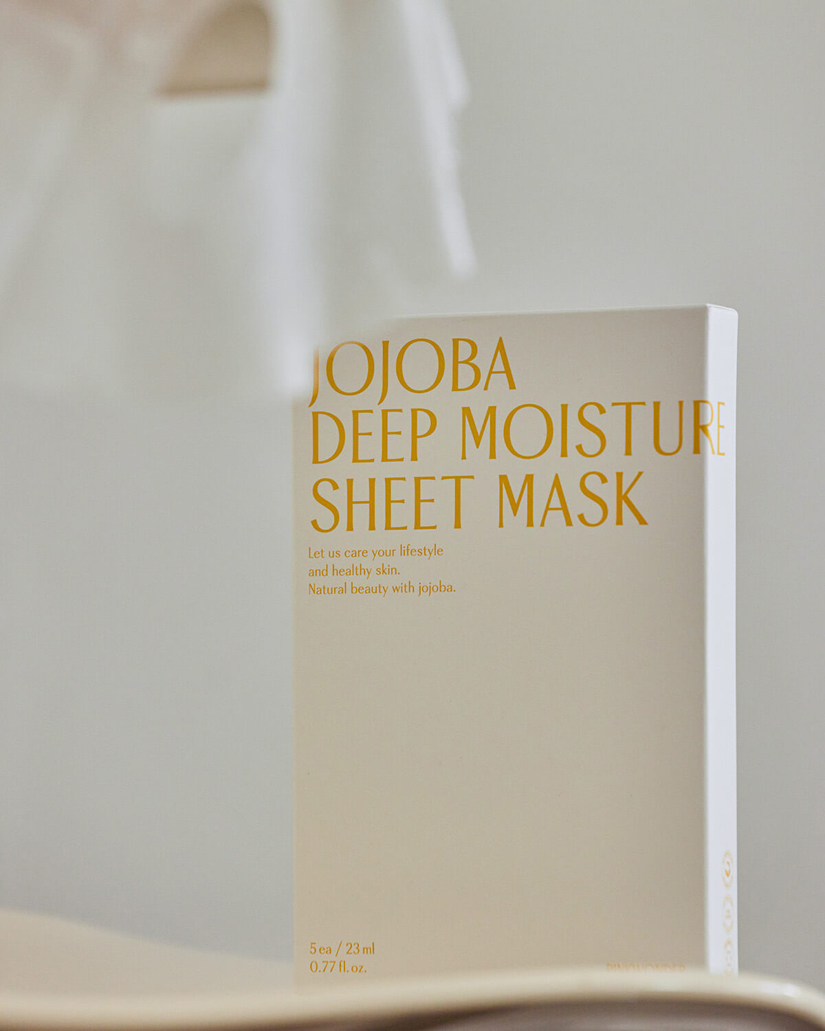 A softly lit, close-up photo of the white box for the Jojoba Deep Moisture Sheet Mask, placed in an indoor setting with a blurred light background. The focus is on the yellow product name and tagline.