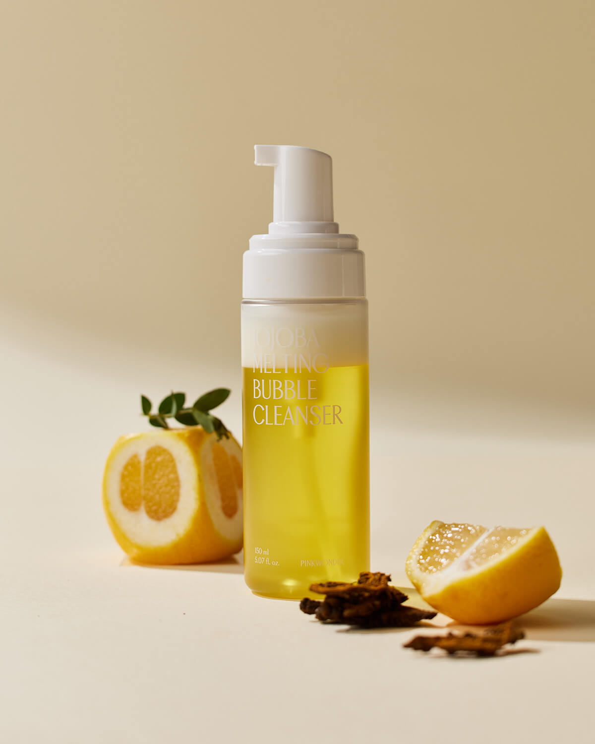 The Jojoba Melting Bubble Cleanser bottle stands upright on a pale tan surface, styled with slices of citrus fruit and dark plant bark, suggesting natural and bright ingredients.