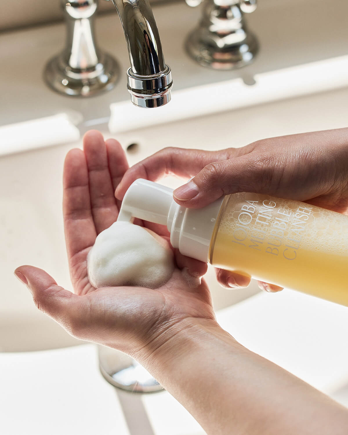 A close-up action shot showing a person's hands dispensing a large, white dollop of Jojoba Melting Bubble Cleanser foam into their palm, with a sink faucet visible in the background.