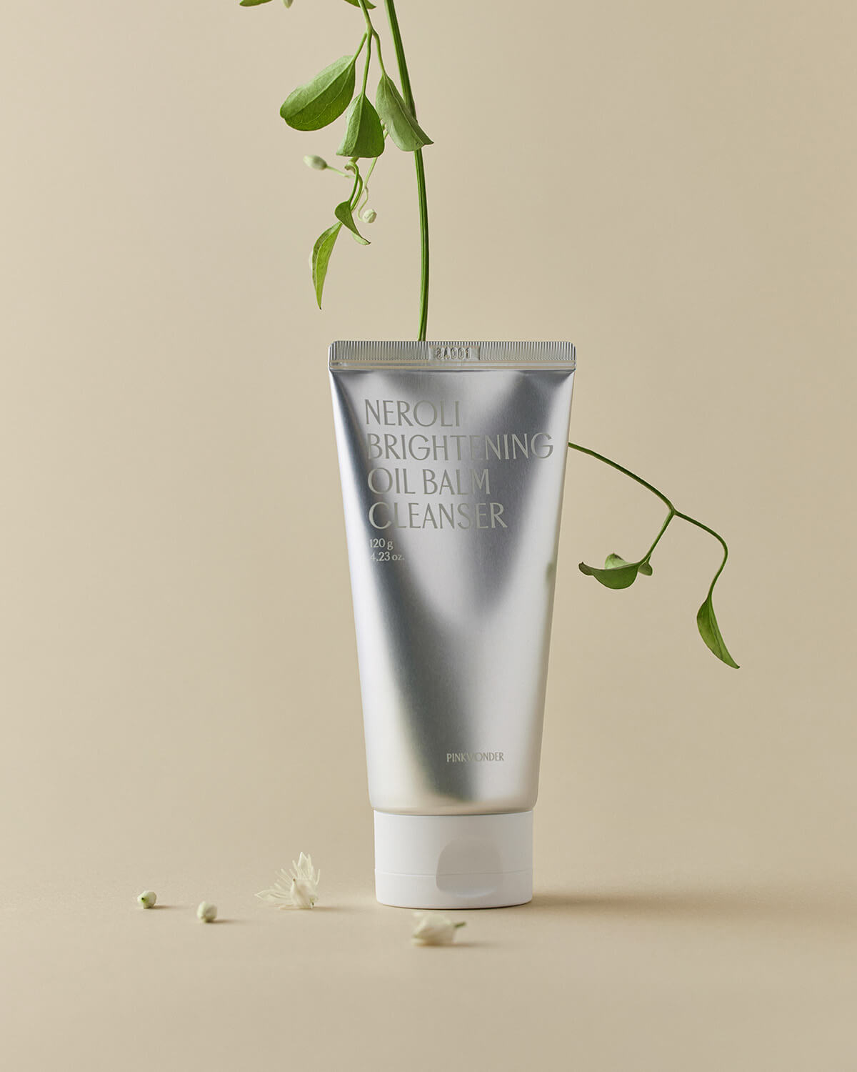 The metallic silver cleanser tube stands upright on a pale tan surface, framed by a delicate vine with small green leaves and white neroli flowers.