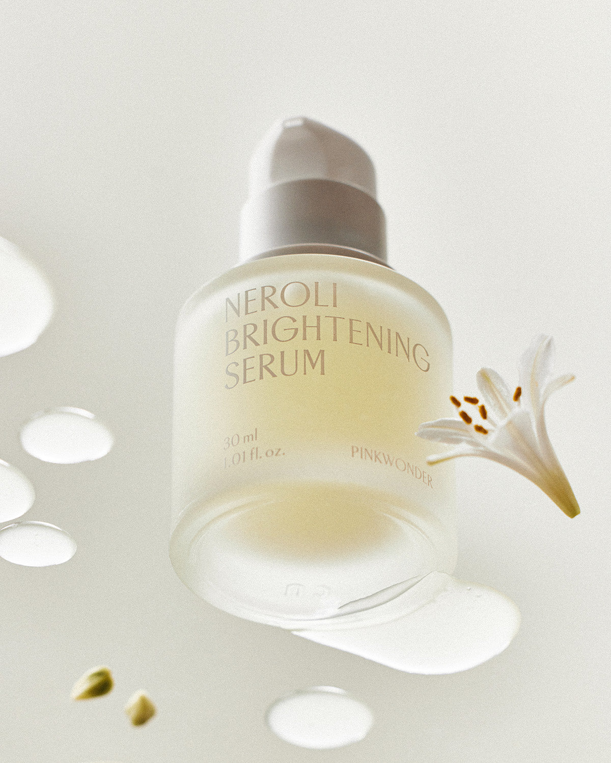 A frosted glass bottle of the serum with a pump top is slightly tilted, surrounded by drops of the serum, two small green seeds, and a delicate white flower with orange stamens.