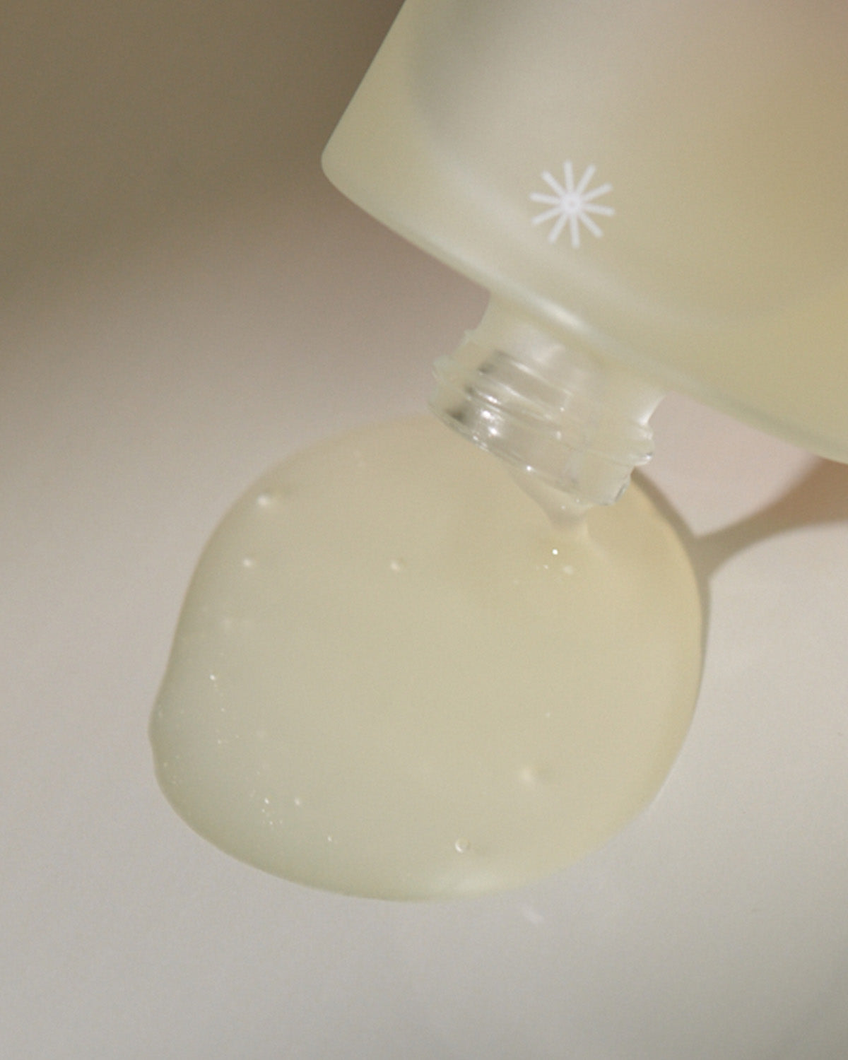 A close-up shot of the light yellow-green serum being poured out of the bottle's neck onto a smooth, pale surface.