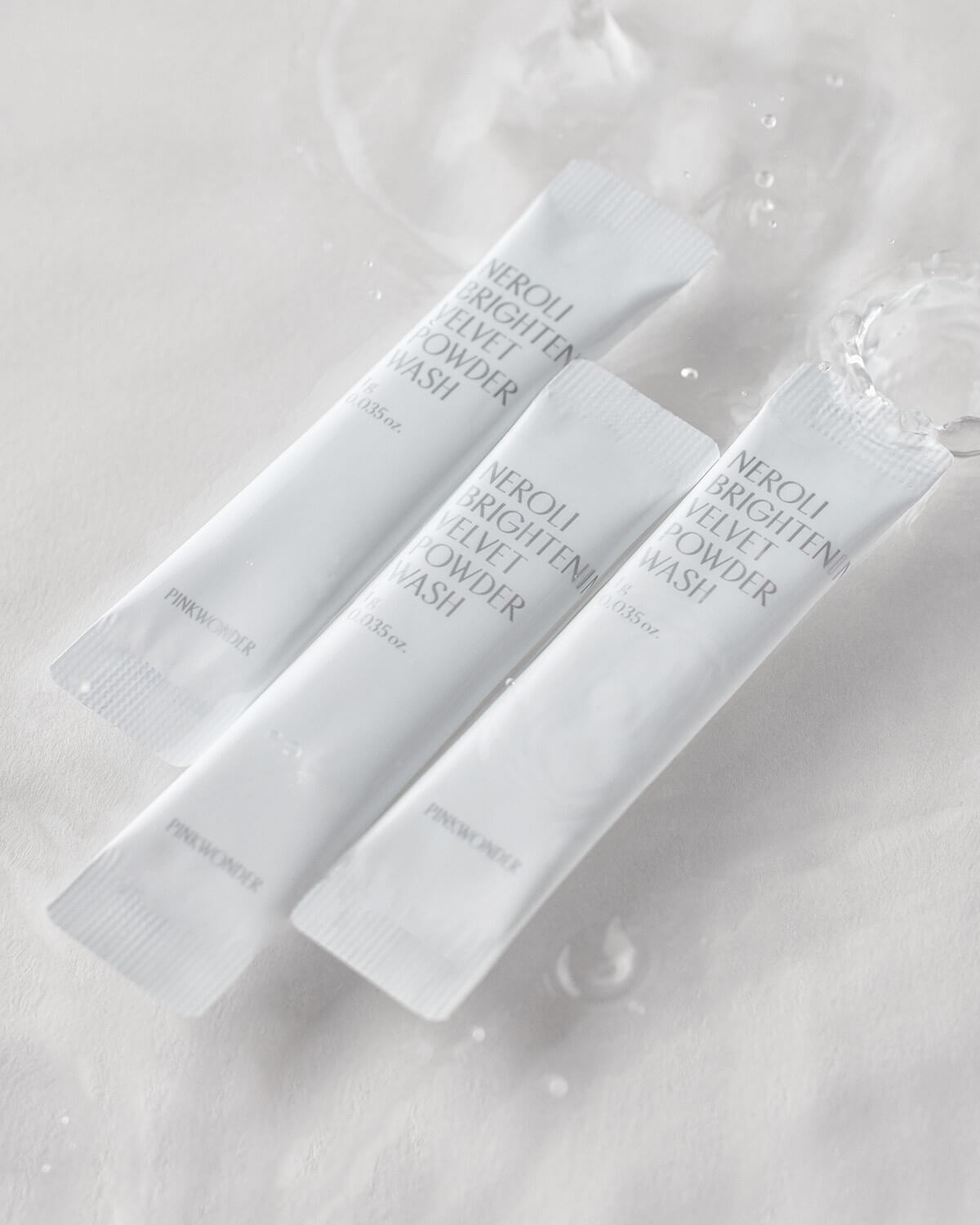 Three white, stick-pack sachets of the "Neroli Brightening Velvet Powder Wash" are arranged diagonally on a white surface, with water splashing and dripping around them.