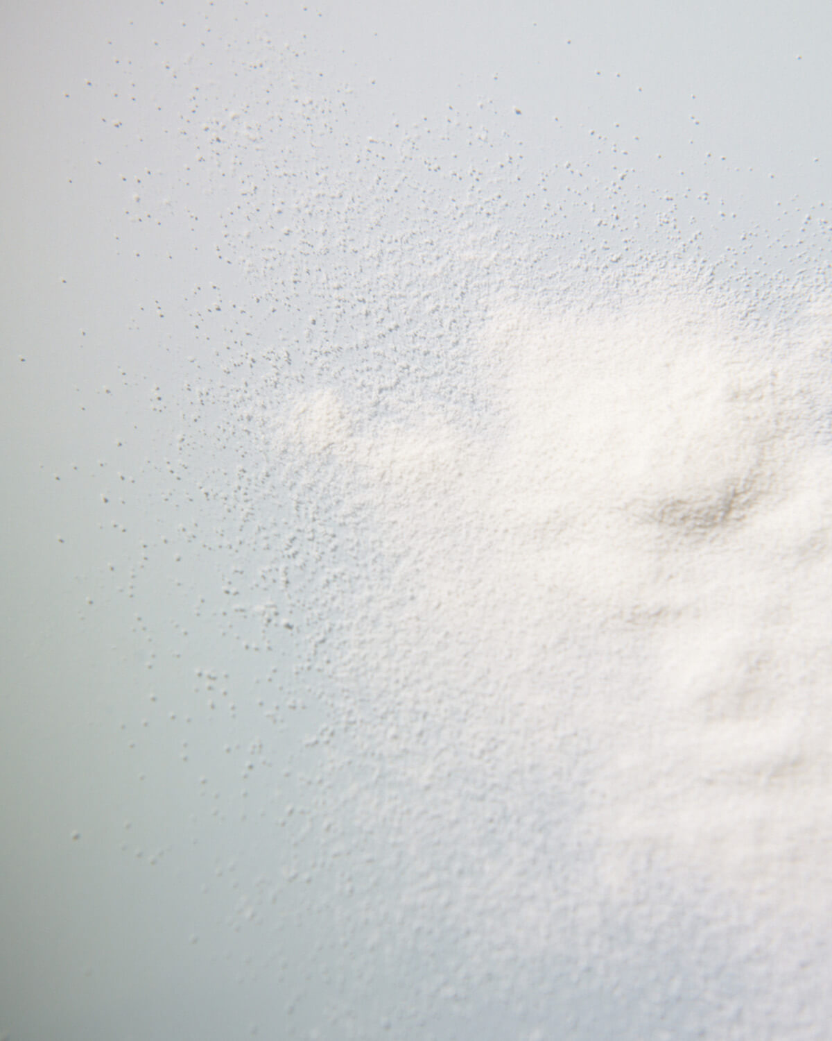 An abstract, high-angle close-up of a pile of fine white powder, presumably the facial wash, scattered across a light blue-gray surface.