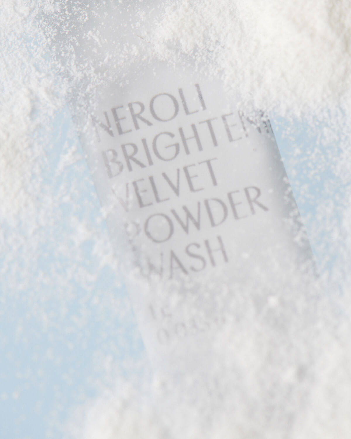 An extreme close-up of a white sachet of "Neroli Brightening Velvet Powder Wash" partially obscured by a cascade of fine white powder, highlighting the product's texture.