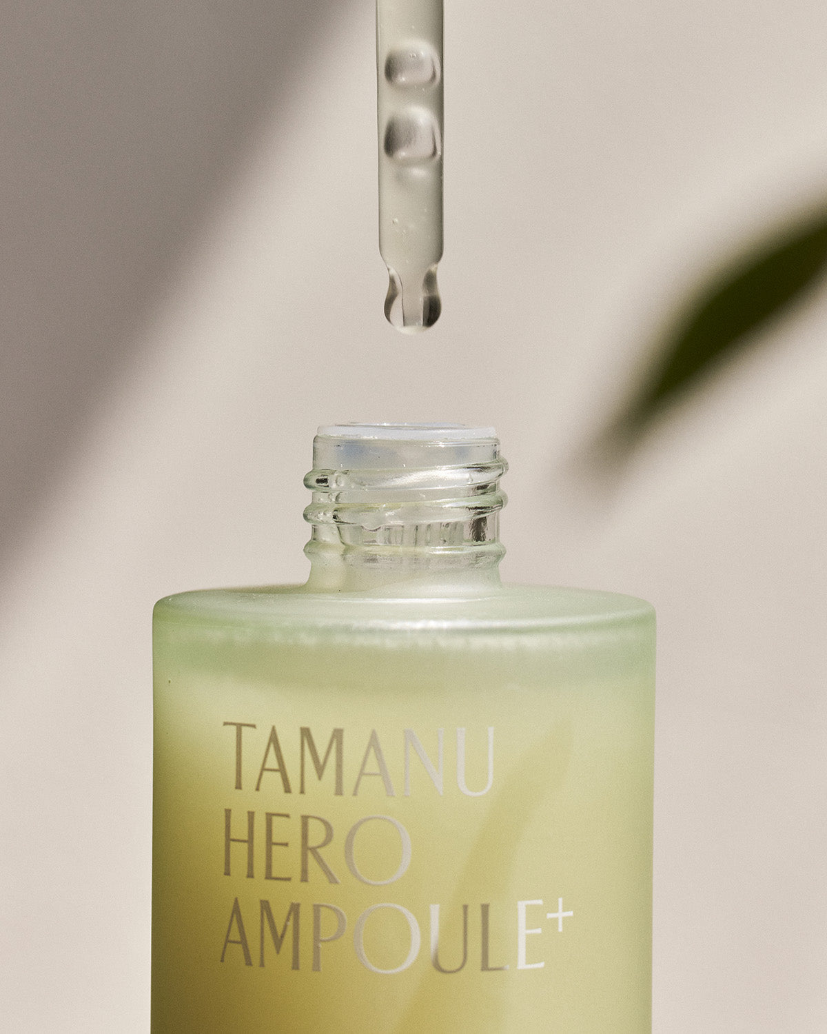 A close-up shot of the Tamanu Hero Ampoule+ bottle. A clear dropper is suspended above the open neck of the bottle, with a bead of serum dripping from its tip.