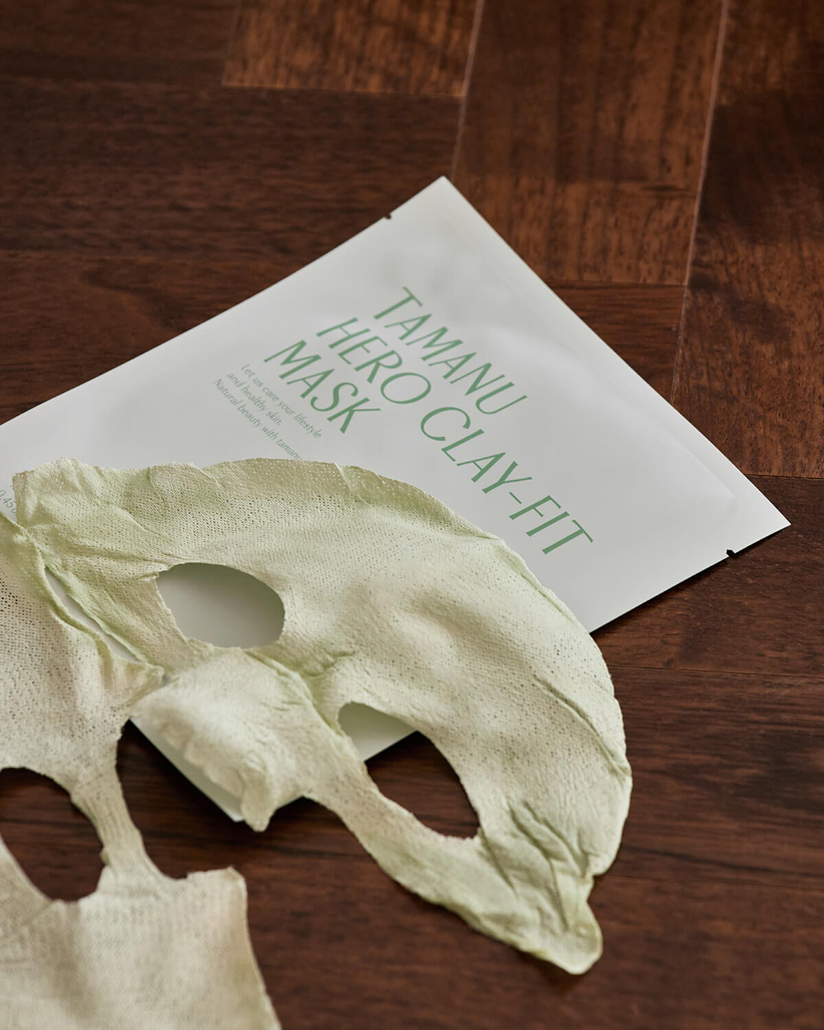 A used, light green, two-piece sheet mask lying on a dark wood floor next to its white and green packaging that reads "Tamanu Hero Clay-Fit Mask."