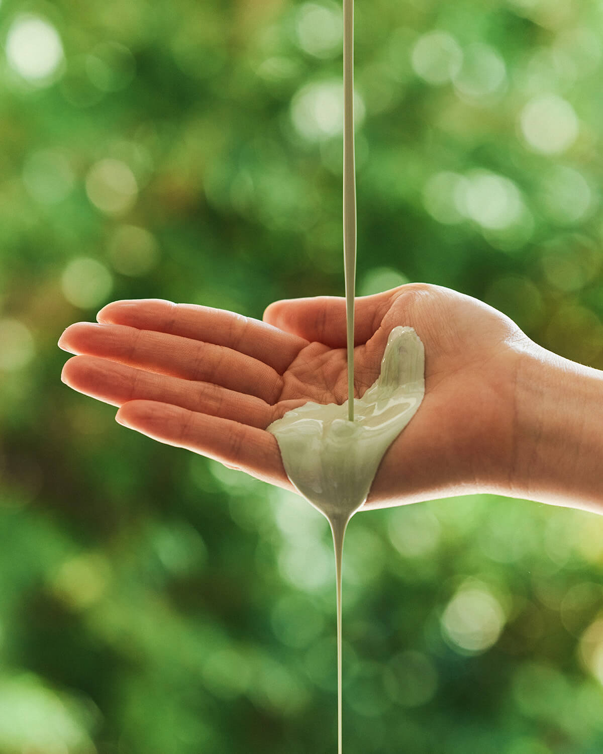 A thick, light green clay product is being poured onto the open palm of a hand. The background is a bright, blurry scene of green trees and nature.
