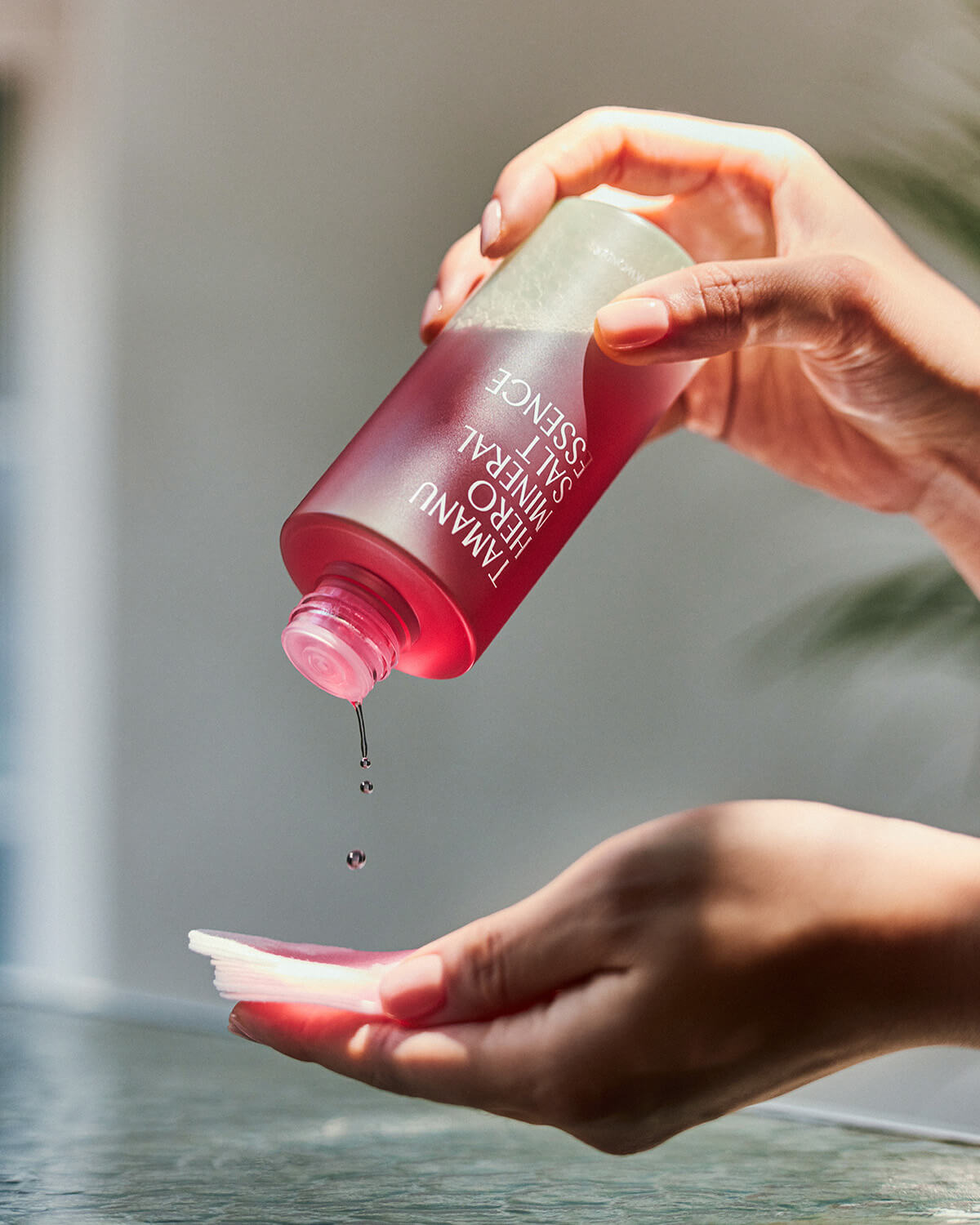 A close-up action shot of a hand holding the bottle of Tamanu Hero Mineral Salt Essence and dispensing drops of the pink-red liquid onto a white cotton pad.