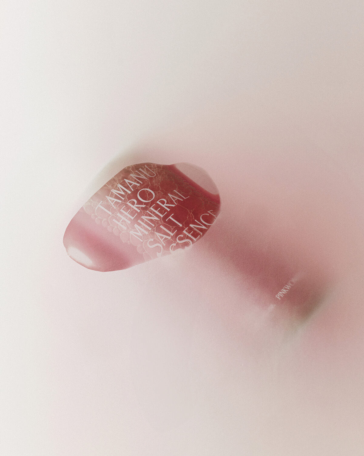 An artistic, blurry shot of the pink-red bottle of Tamanu Hero Mineral Salt Essence lying on a surface, viewed through a soft, pink-tinted filter.