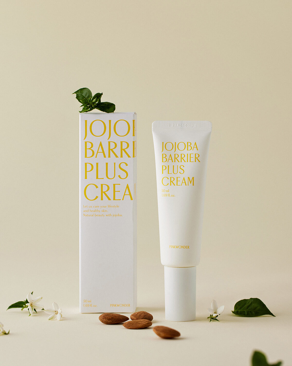 The white product box and the matching tube of Pinkwonder Jojoba Barrier Plus Cream are shown standing upright on a pale tan background. They are styled with several almonds, small white flowers, and green leaves to emphasize the natural ingredients.