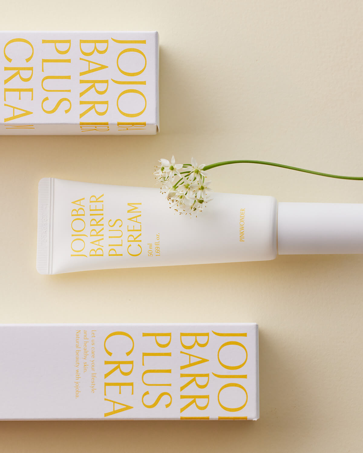 A flat-lay photo of the Pinkwonder Jojoba Barrier Plus Cream tube, its cap to the right, placed horizontally between two white boxes of the same product. A delicate white flower with a thin green stem is draped over the top of the tube, all set on a light tan surface.