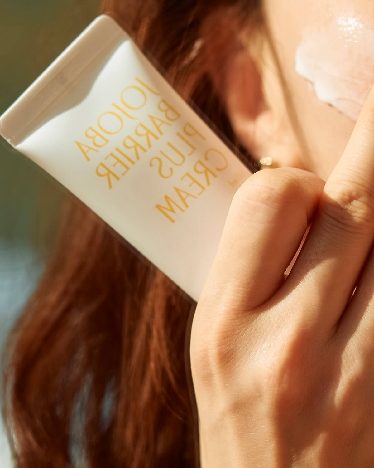 A close-up action shot of a person applying the cream to their face. A hand holds the Pinkwonder Jojoba Barrier Plus Cream tube, with the reflection of the product name visible, as they use a finger to blend the white cream onto their cheek.