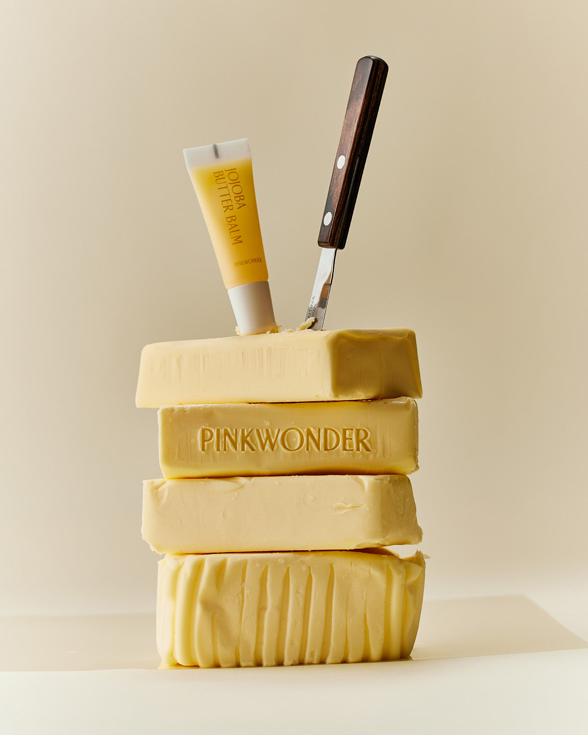 An artistic product photo featuring a tall stack of four sticks of pale yellow butter. The second stick from the top is embossed with the word "PINKWONDER." A small yellow tube of Jojoba Butter Balm and a butter knife are stuck into the top stick of butter.