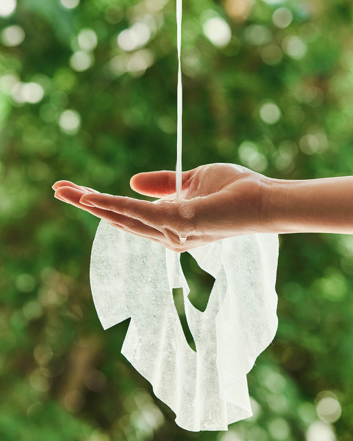 A close-up outdoor shot of a hand holding a saturated white sheet mask. A stream of clear liquid, representing the serum, is pouring onto the hand and the mask below, with a blurred green natural background.