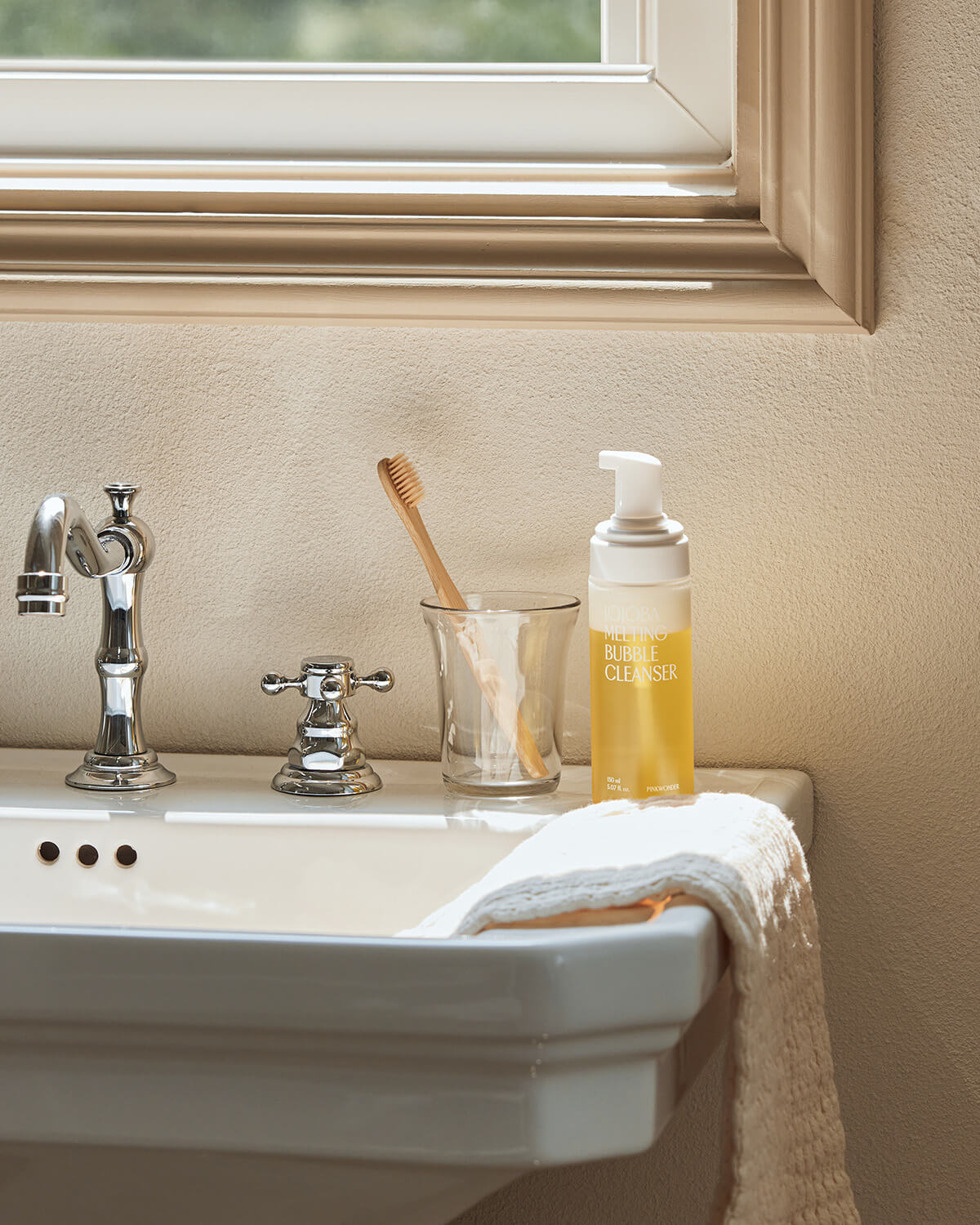 The Jojoba Melting Bubble Cleanser bottle sits on the edge of a bathroom sink next to a toothbrush in a glass and chrome fixtures. The background features a window with natural light filtering in.