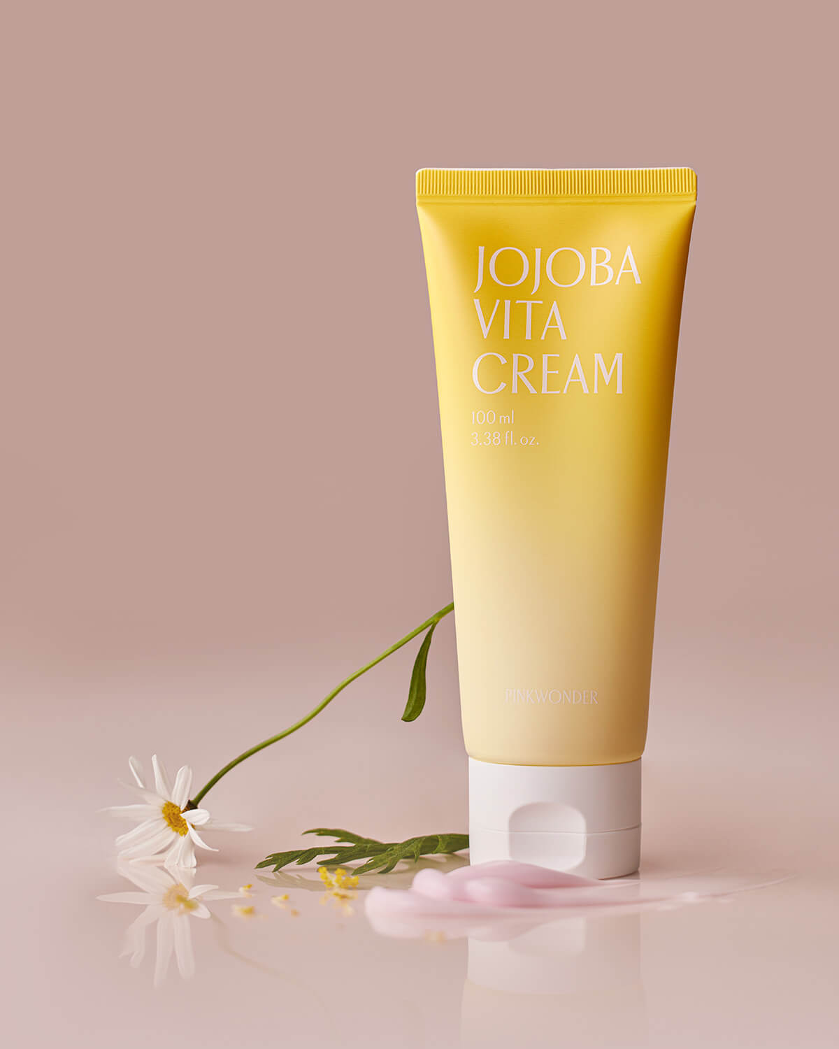 An artistic product shot of the yellow Jojoba Vita Cream tube standing upright on a reflective pink surface. A dab of pink cream is squeezed out near the white cap, and a white daisy is positioned on the left.