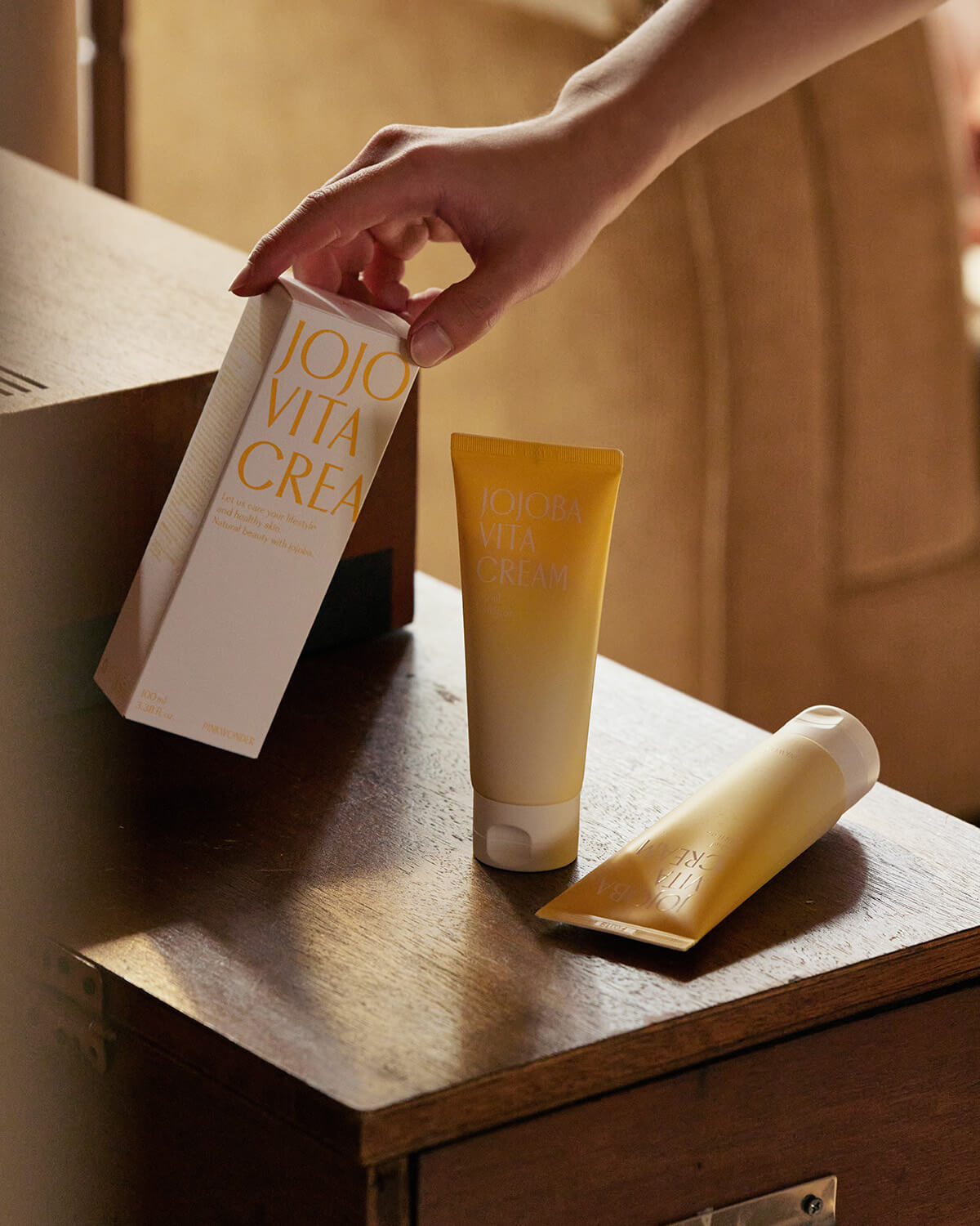 A warm, indoor scene with a hand reaching down to place the white box of Jojoba Vita Cream next to two tubes of the product on a wooden bedside table. The room has soft, inviting lighting.
