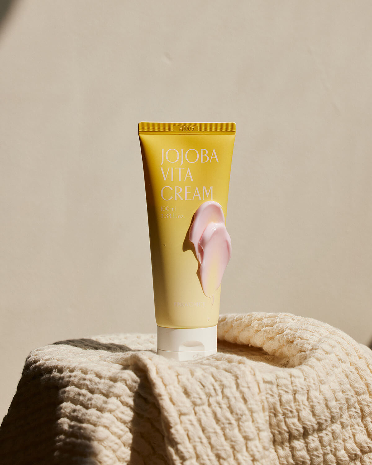 The yellow Jojoba Vita Cream tube stands on a light beige knitted sweater or towel, set against a smooth, bright background. A thick, wavy dollop of pink cream is artfully placed on the front of the tube.