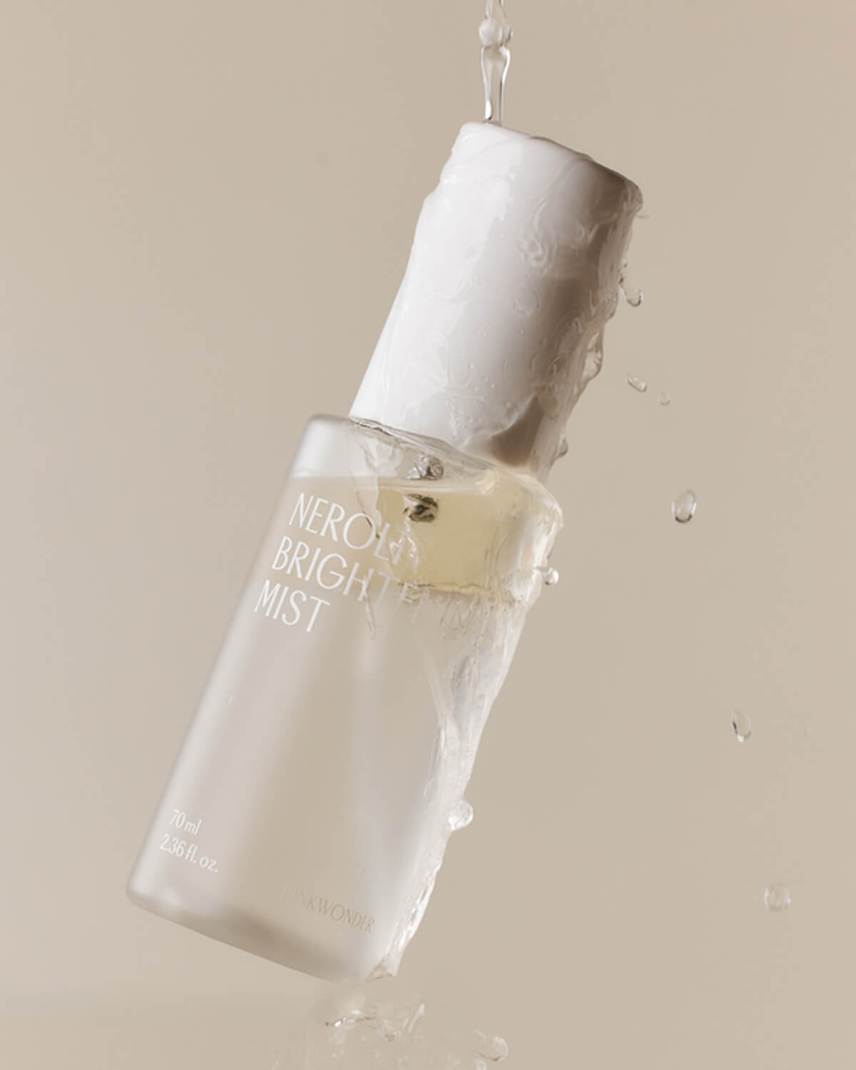 A product shot of the mist bottle tilted on a light tan background, with water dramatically pouring and splashing over the cap and the top portion of the bottle.