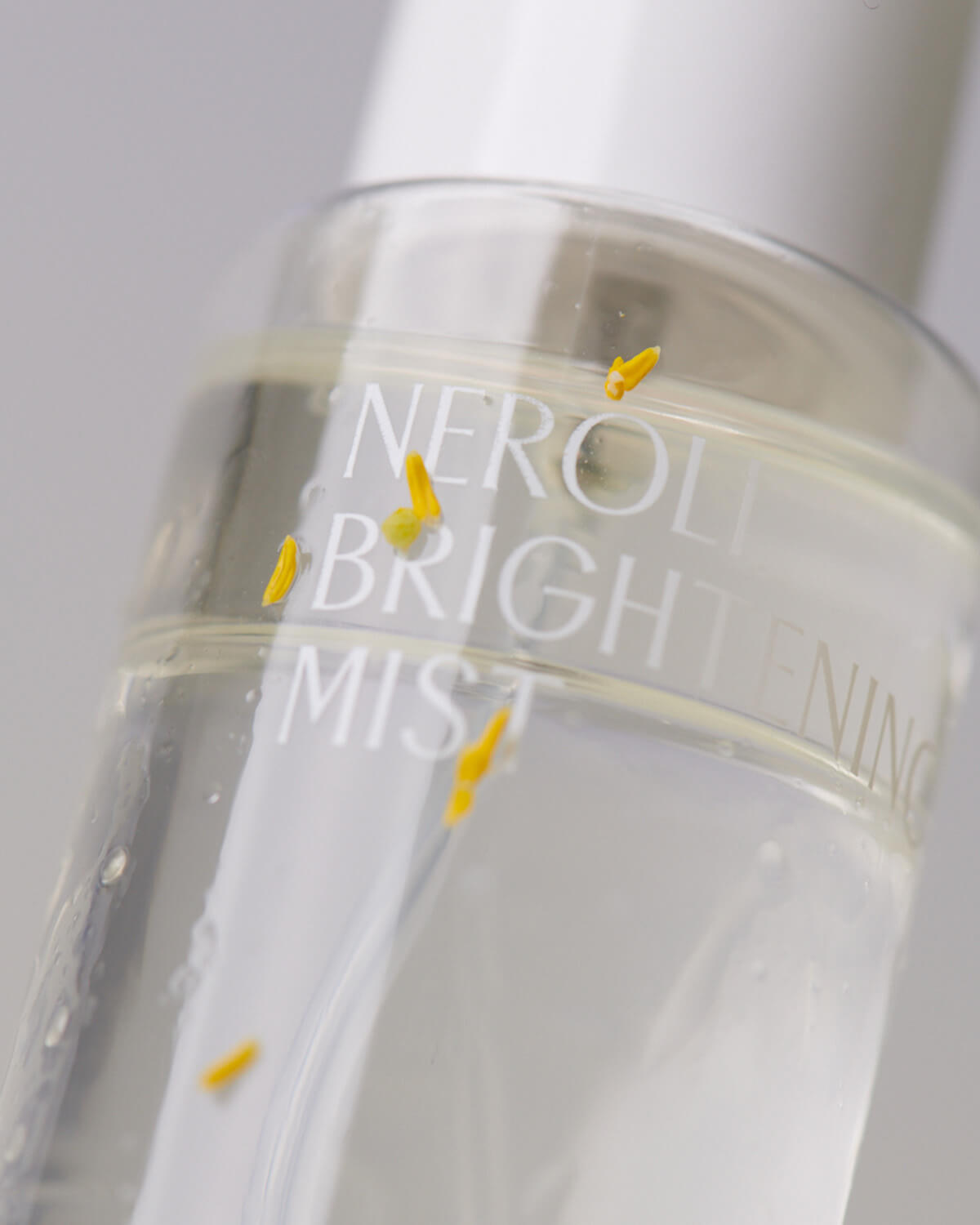 An extreme close-up of the clear mist bottle showing the liquid inside, which contains small, bright yellow particles or petals floating near the "Neroli Brightening Mist" text.