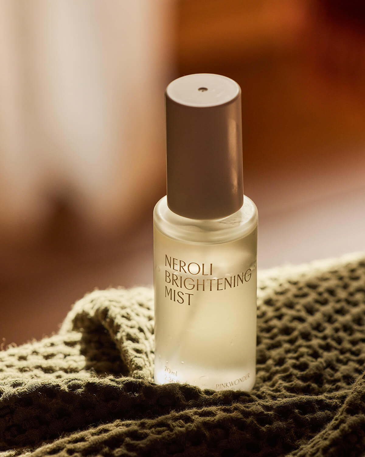 A close-up, dramatic shot of the mist bottle resting on a textured, olive green knitted fabric. The background is a soft, blurry mix of dark and light browns.