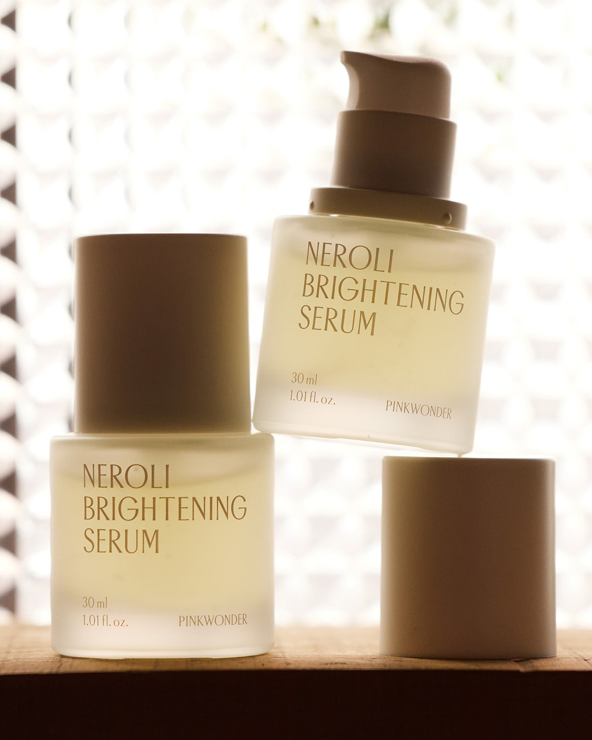 Two frosted glass bottles of "Neroli Brightening Serum" by Pinkwonder, one with a pump dispenser and the other with a cap, are creatively stacked against a bright, patterned background.