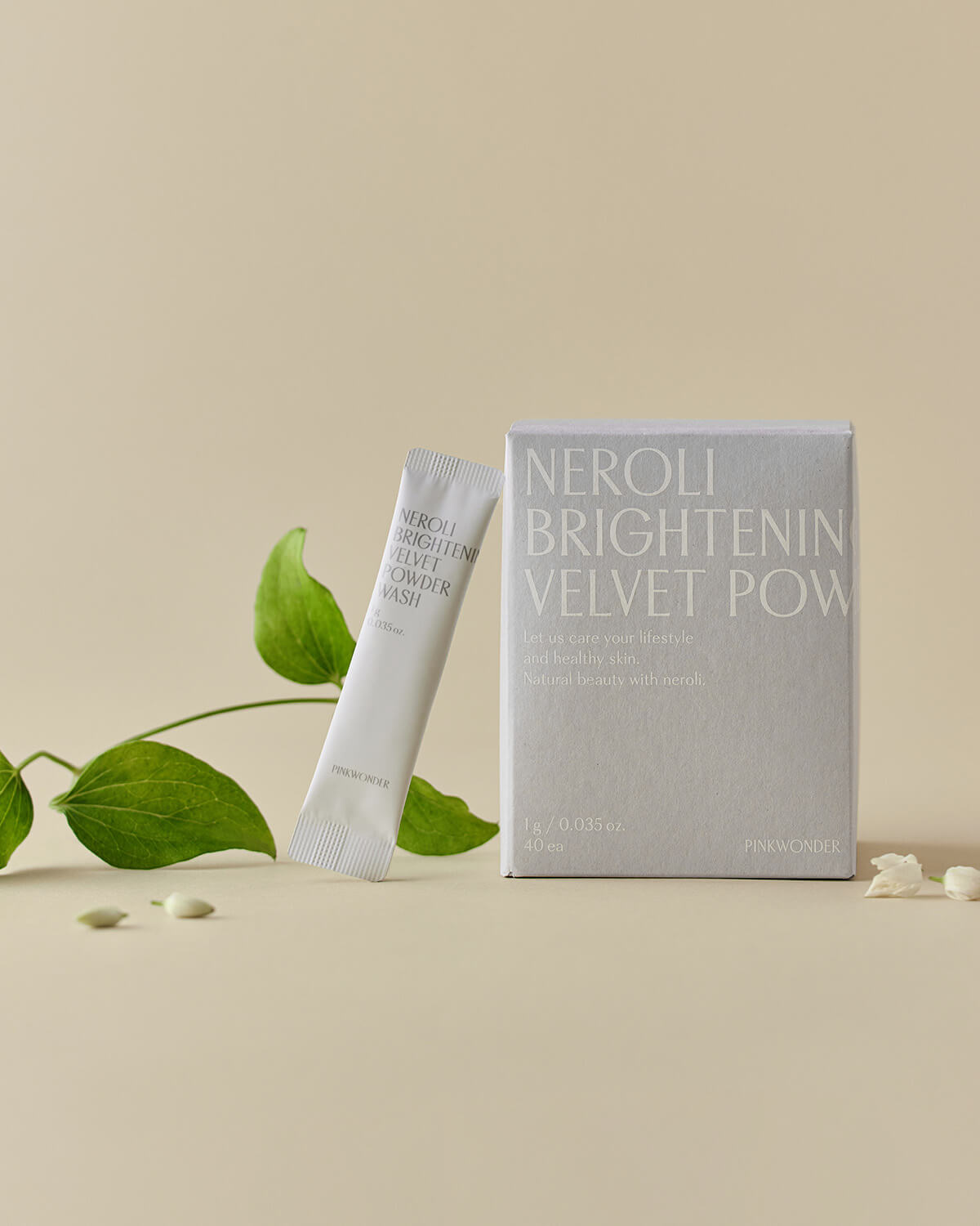 An artistic product composition featuring the light gray box and a single sachet of the powder wash, arranged with a sprig of greenery, small seeds, and tiny white flowers against a pale tan background.