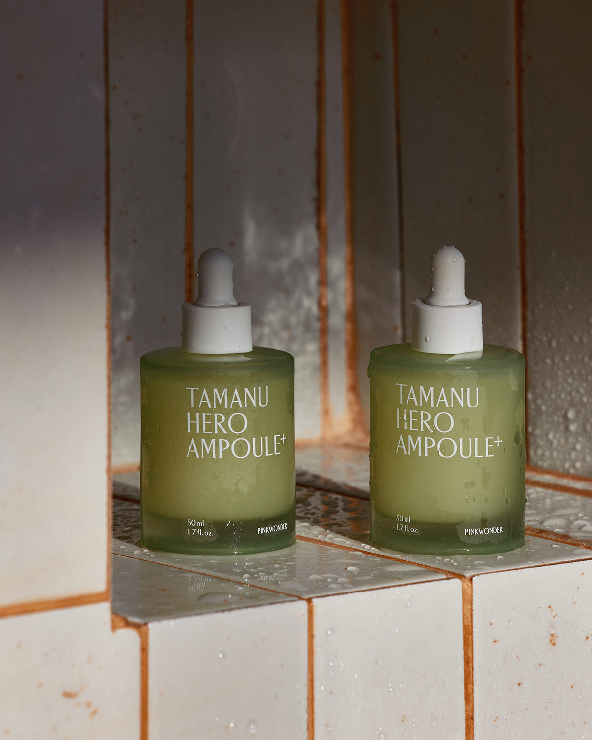 Two identical bottles of the frosted glass Tamanu Hero Ampoule+ stand on a tiled shelf, sprinkled with water drops, under a warm light.