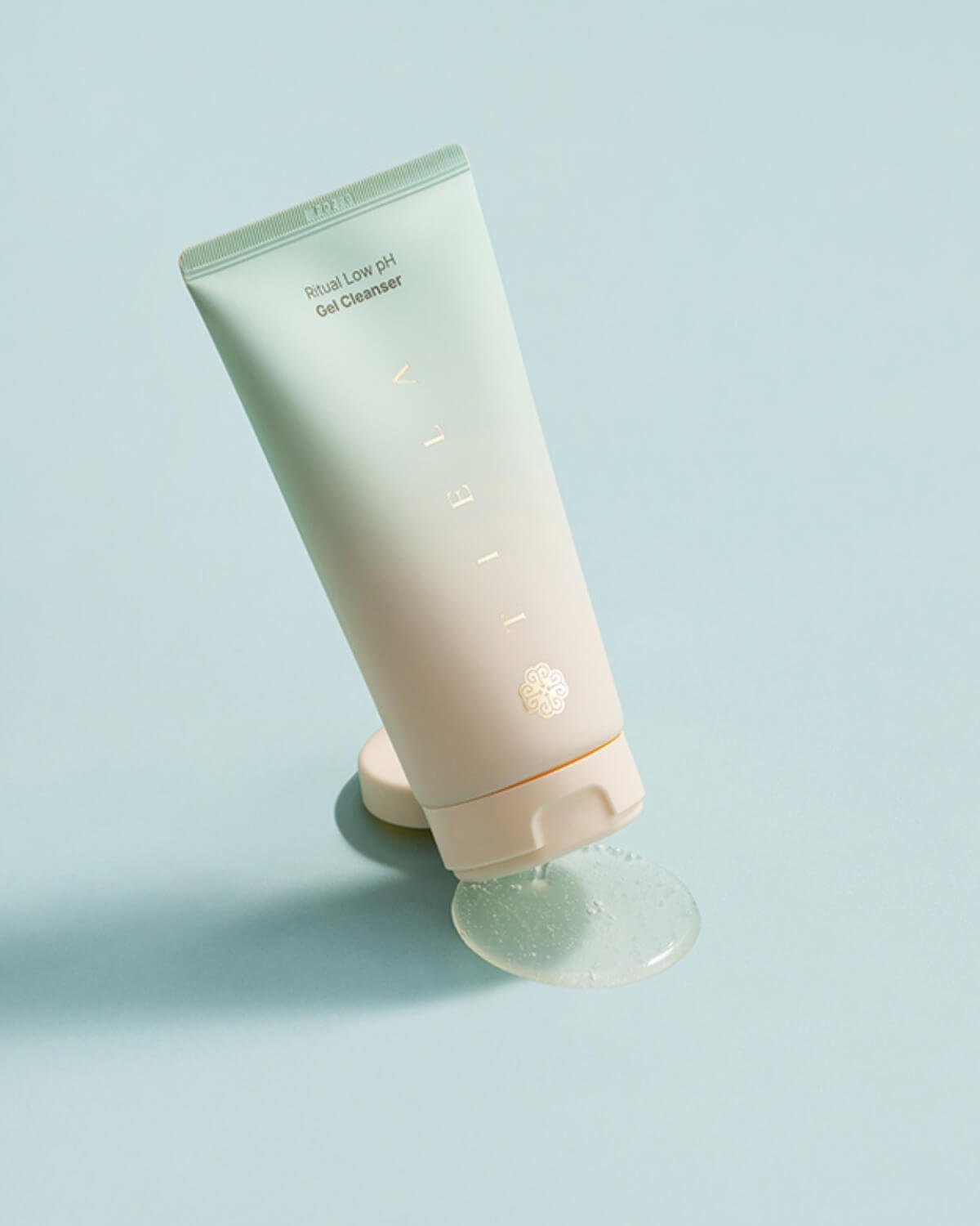 The Tiela Low pH Gel Cleanser tube is tilted on a light blue surface, with a puddle of the clear gel cleanser product dripping out of the opening.