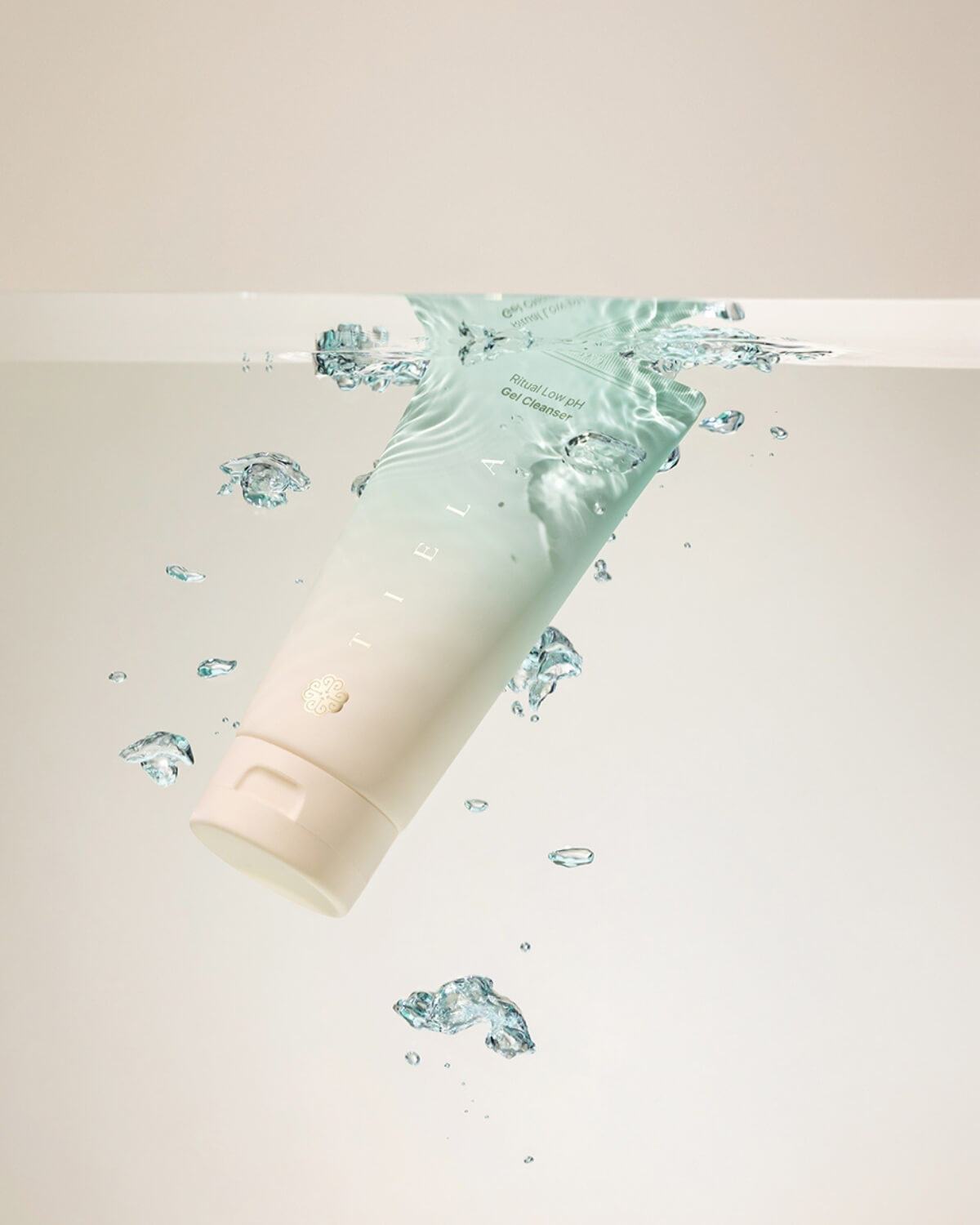 The Tiela Low pH Gel Cleanser tube is being submerged into water, creating bubbles and splashes around the product.
