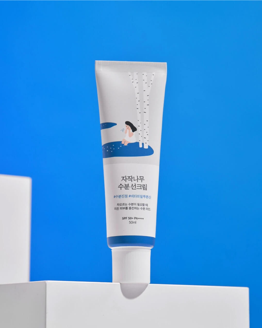 The full tube of Round Lab Birch Moisturizing Sunscreen is presented vertically on white platform blocks against a striking blue background, highlighting the Korean text and SPF 50+ PA++++ rating on the packaging.