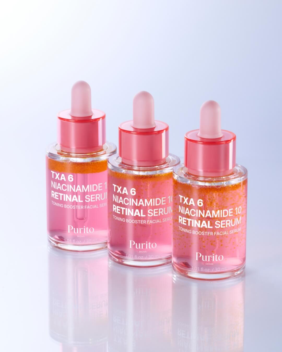 A line of three dropper bottles of Purito TXA 6 Niacinamide 10 Retinal Serum, showcasing the product's pink and orange bi-phase formula and reflective glass bottles on a subtly gradient white background.