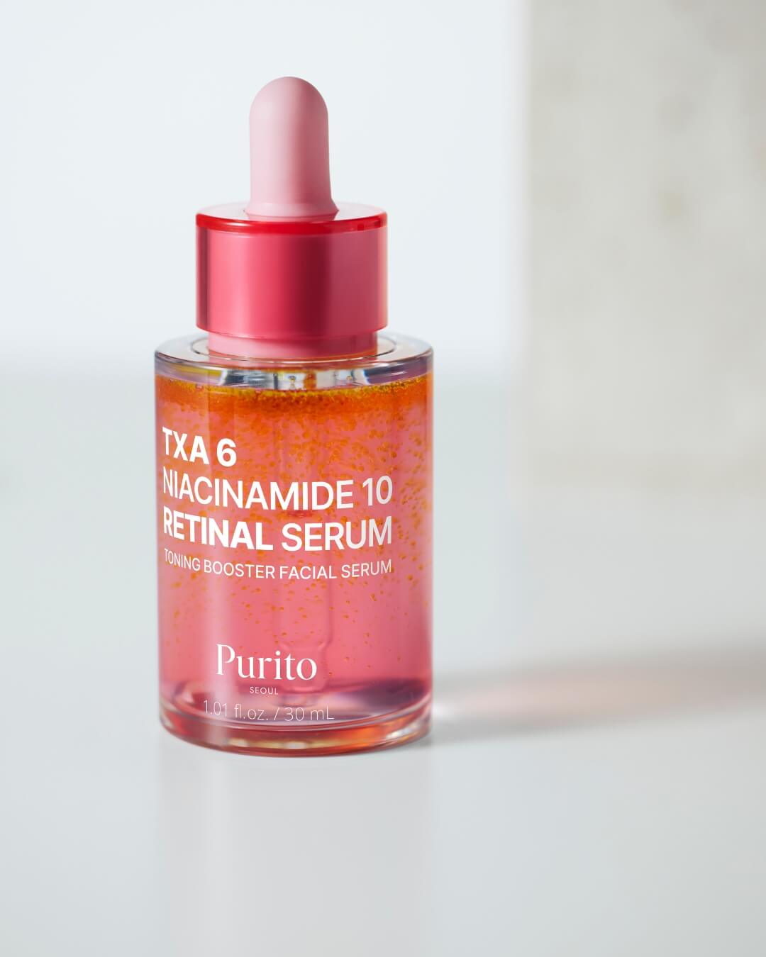 A close-up, slightly soft-focus studio image of a closed bottle of Purito TXA 6 Niacinamide 10 Retinal Serum. The bi-phase mixture, which must be shaken to combine, is clearly separated into a bright orange top layer and a pink bottom layer with orange specks.