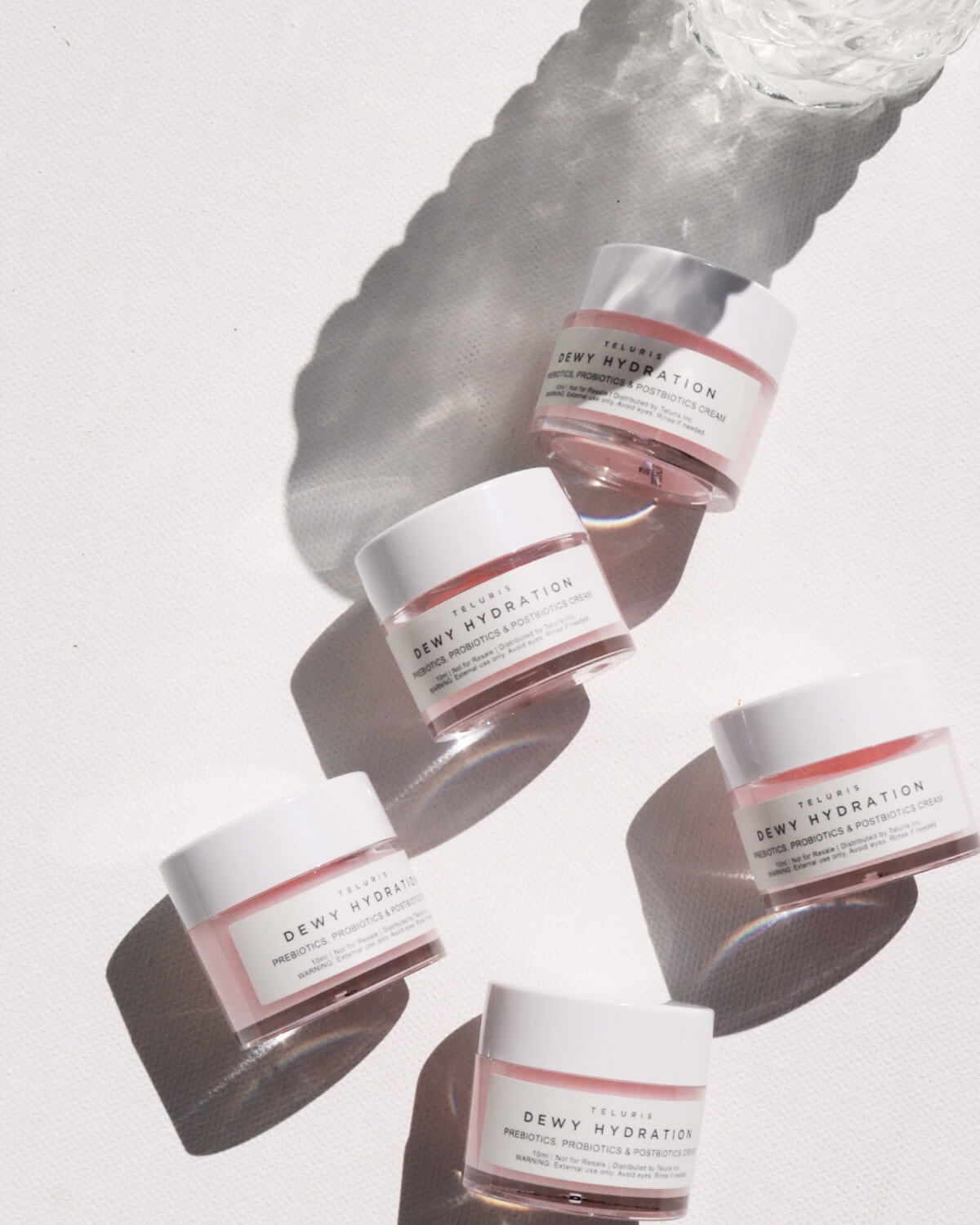 Five small jars of Teluris Dewy Hydration cream with pink product and white lids under bright sunlight.
