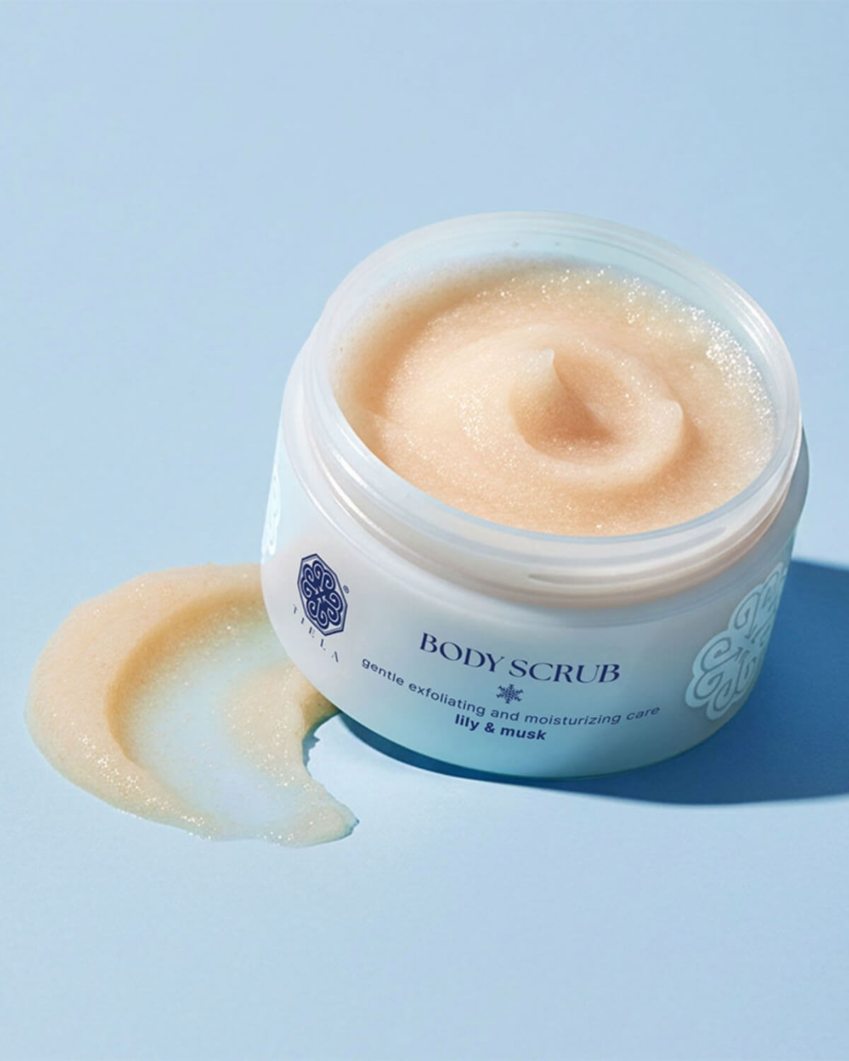 An open jar of Tiela Lily & Musk body scrub showing its grainy texture and a small smear of the product.