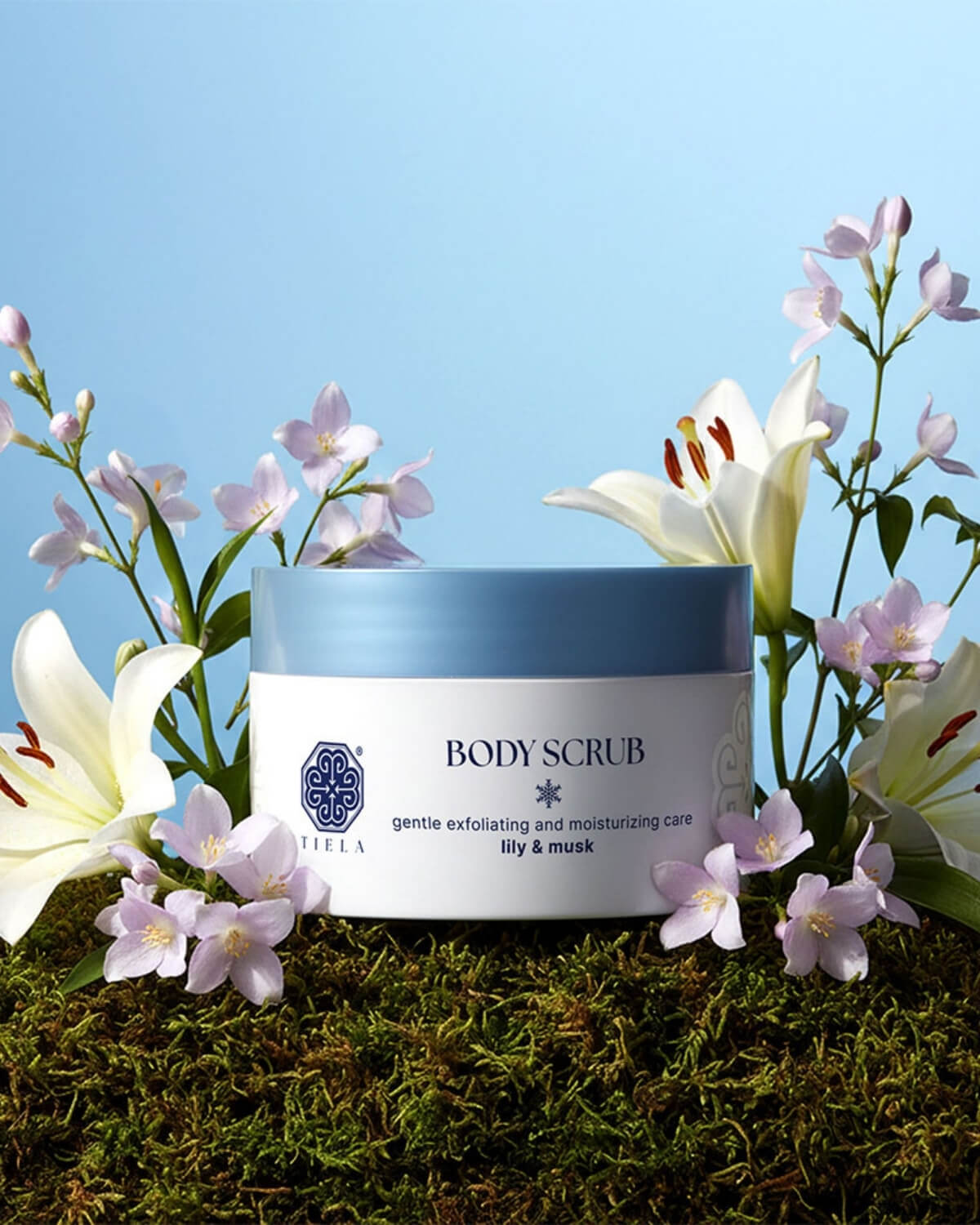 A jar of Tiela Lily & Musk body scrub centered among moss and small purple and white flowers on a blue backdrop.