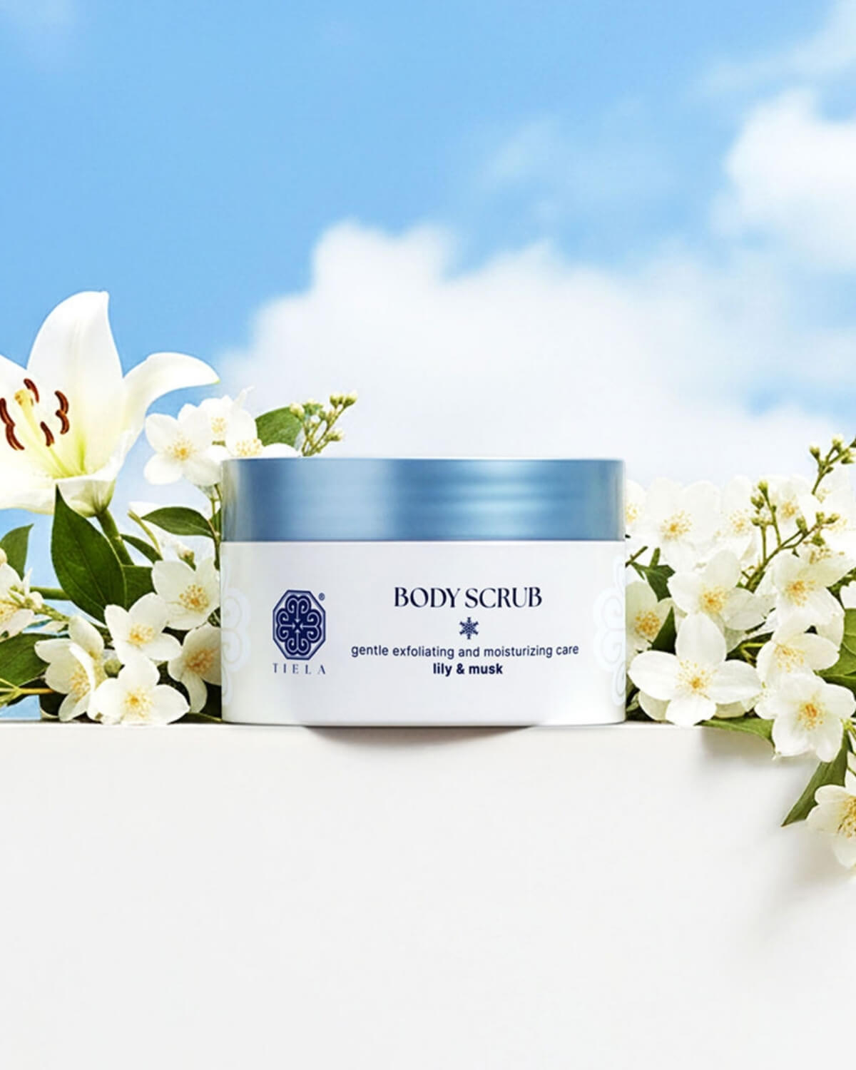 Tiela Lily & Musk body scrub jar framed by white flowers against a bright blue sky with white clouds.
