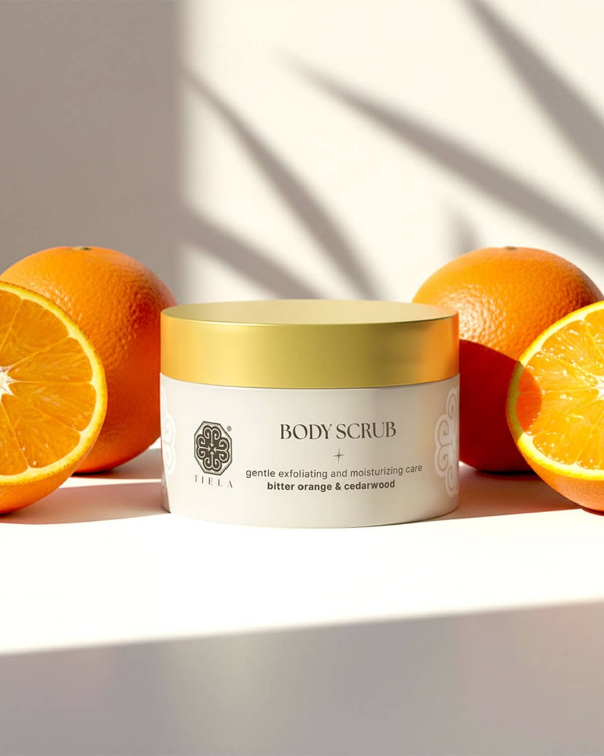 Tiela Bitter Orange & Cedarwood body scrub jar surrounded by whole and halved oranges under bright sunlight.