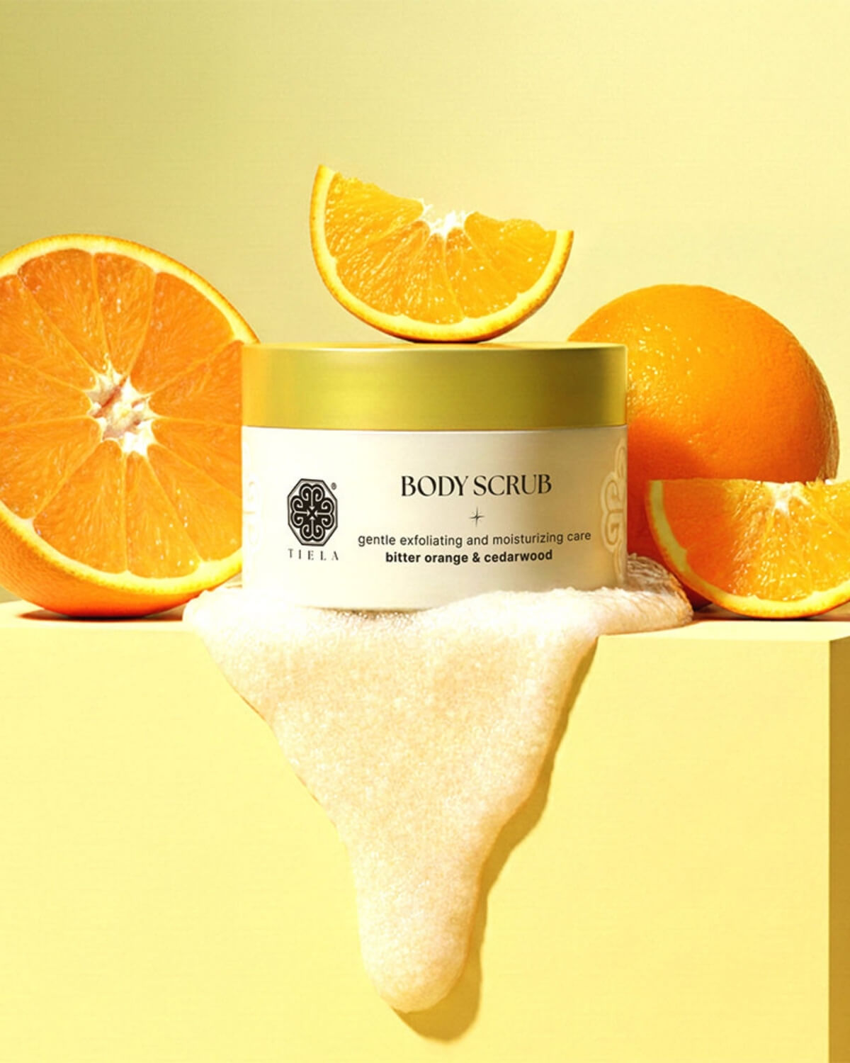 Tiela body scrub jar on a yellow pedestal with orange slices and a thick product drip cascading down the front.