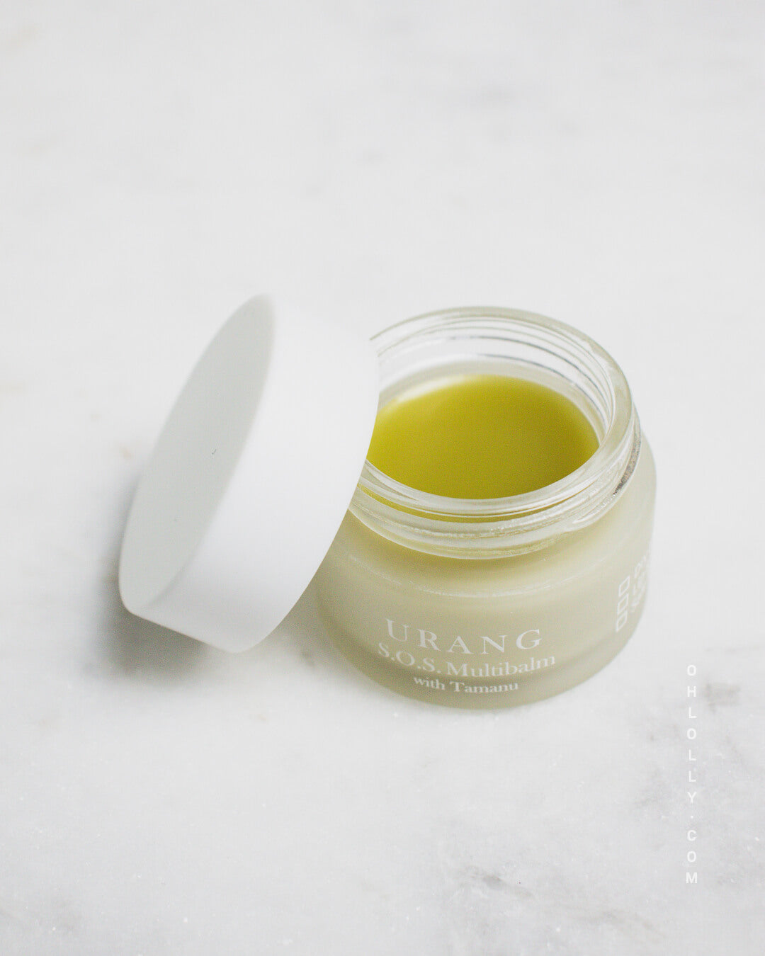 Close-up of the Urang S.O.S. Multi Balm with Tamanu in a small, clear glass jar with a white lid slightly ajar, showing the solid, pale yellow-green balm inside on a white marble surface.