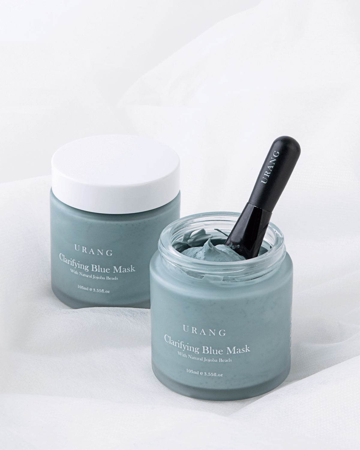 Two jars of Urang Clarifying Blue Mask, one open with an application brush dipped in, illustrating the soft, easily spreading clay and gentle exfoliation.