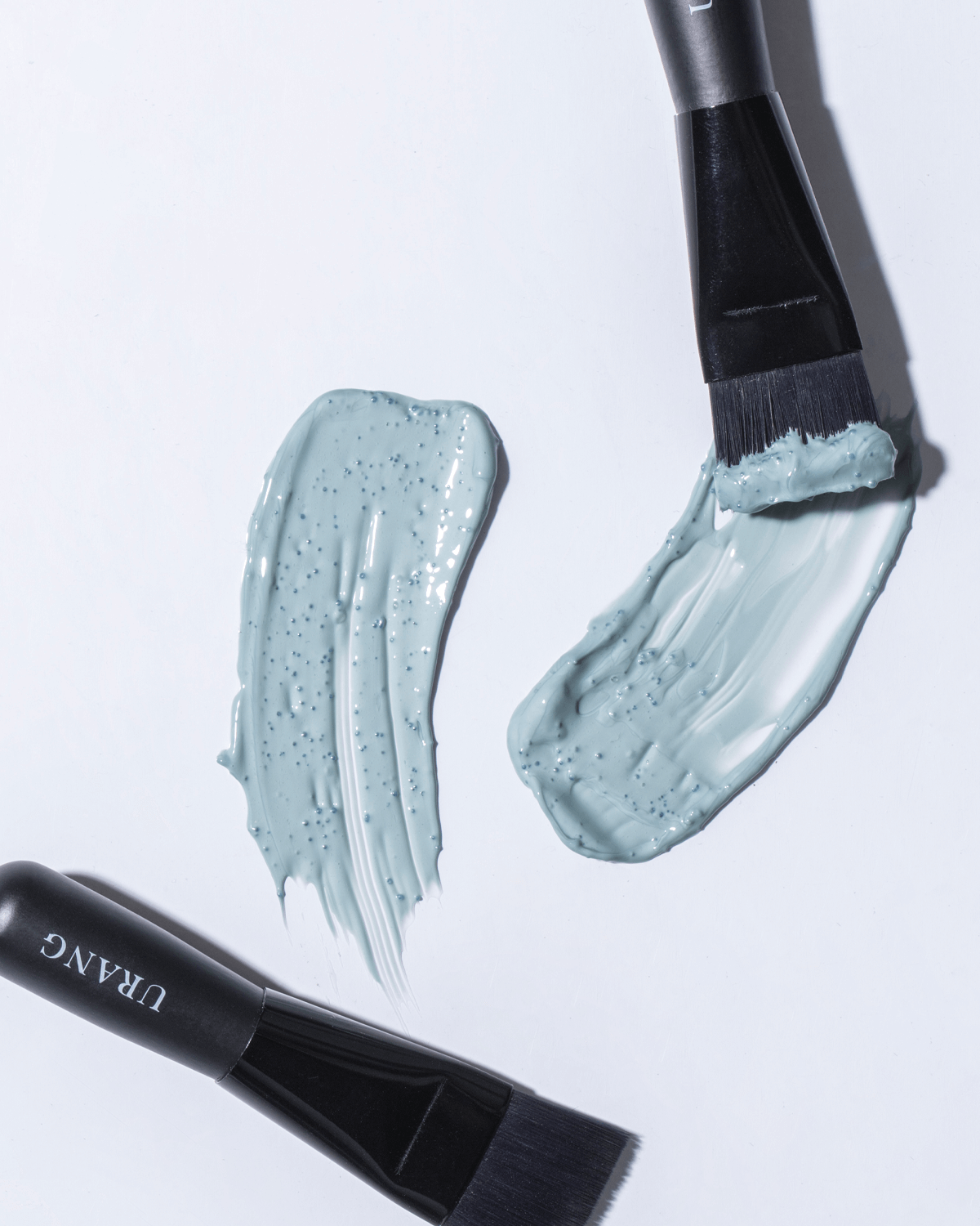 Close-up showing swatches of the soft blue Urang Clarifying Blue Mask next to two brushes, illustrating the creamy texture and gentle exfoliation from jojoba beads.
