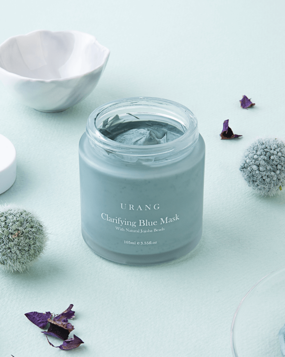 Urang Clarifying Blue Mask jar on a serene light blue backdrop with decorative florals and a white bowl emphasizing its organic, vegan ingredients and gentle nature.
