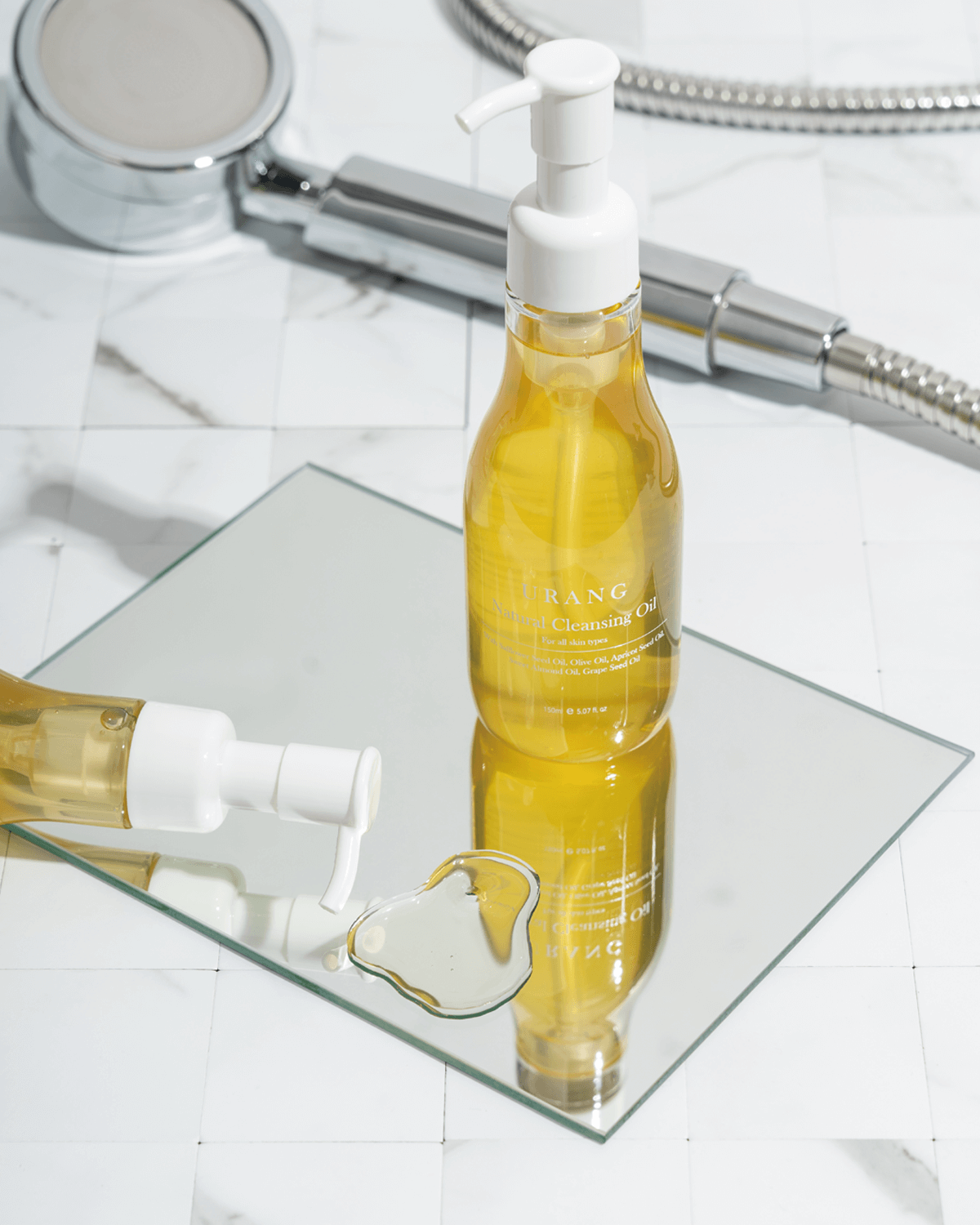 The clear glass pump bottle of Urang Natural Cleansing Oil standing on a mirror, with a puddle of the golden-yellow oil spilled onto the surface and a metallic shower head in the background.