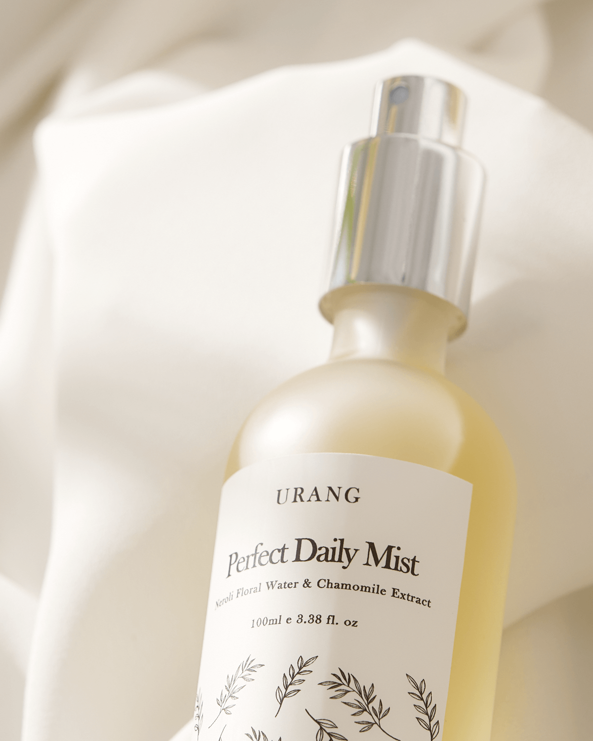 Close-up of the Urang Perfect Daily Mist frosted glass bottle, highlighting the minimalist white label with black text and the silver spray cap against a soft, light-colored fabric background.
